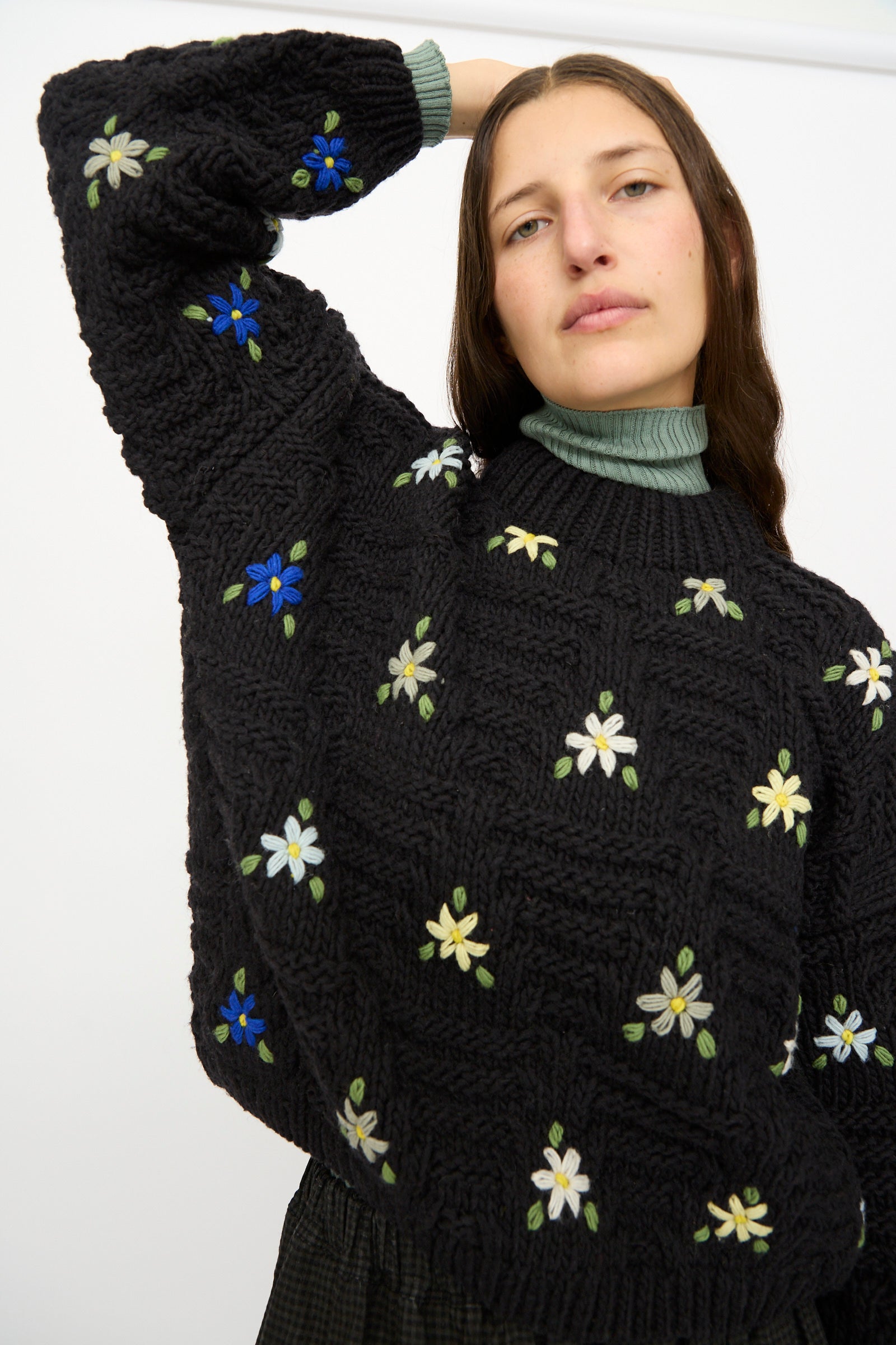 A person wears the Ichi Antiquités Wool Hand-knit Pullover with Flowers in Black, featuring colorful embroidered flowers, with one arm raised behind their head.