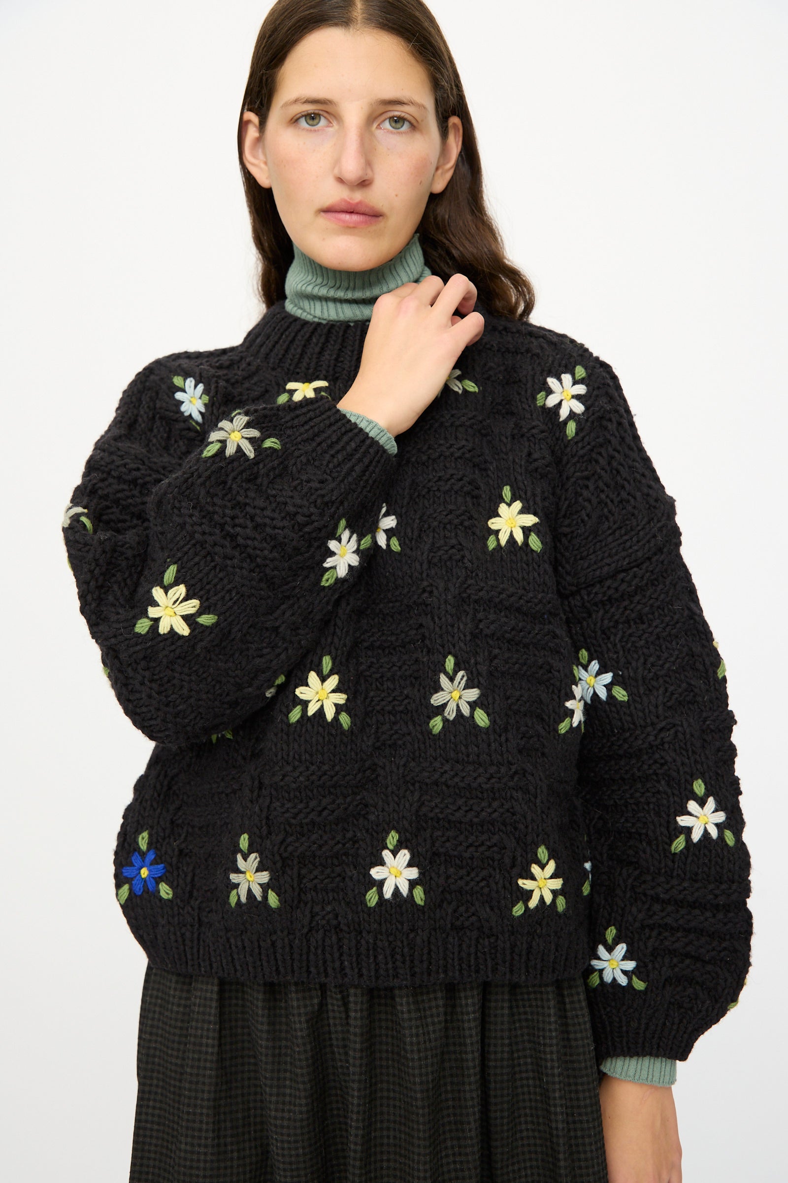 A woman with long brown hair models the Ichi Antiquités Wool Hand-knit Pullover with Flowers in Black, paired with a green turtleneck and dark skirt, standing against a plain white background.