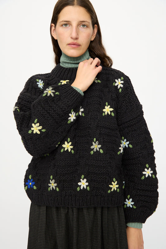 A woman with long brown hair models the Ichi Antiquités Wool Hand-knit Pullover with Flowers in Black, paired with a green turtleneck and dark skirt, standing against a plain white background.