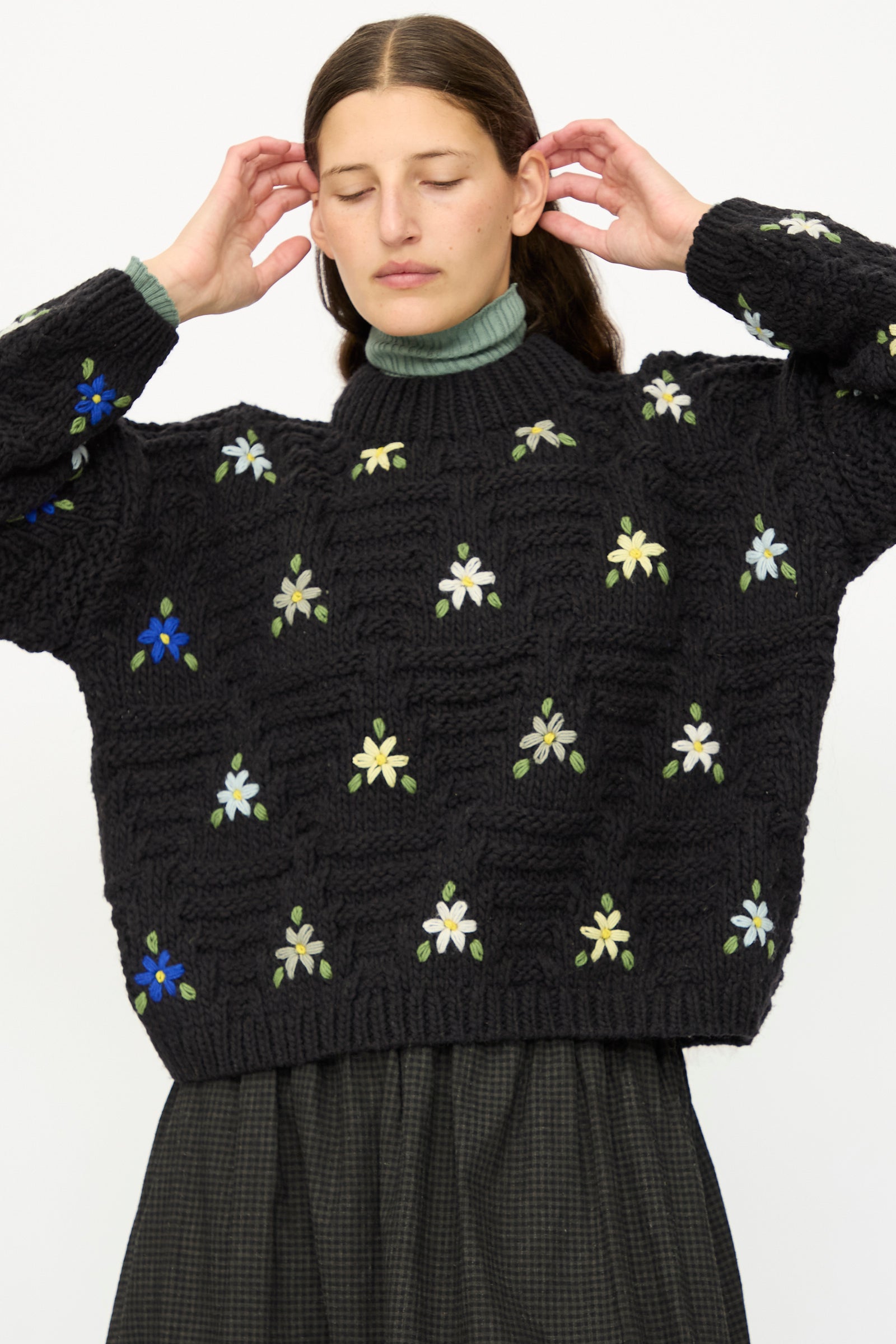 A woman stands with eyes closed, wearing the Ichi Antiquités Wool Hand-knit Pullover with Flowers in Black over a green turtleneck and a dark checkered skirt, her hands raised near her head.