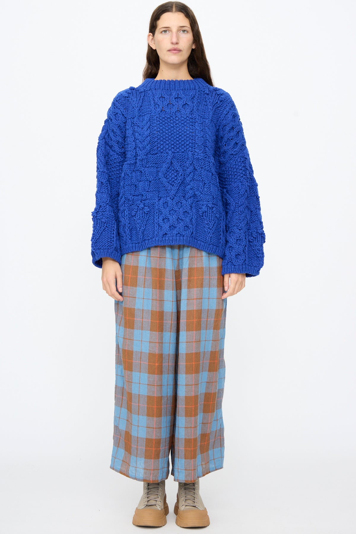 A person wearing an Ichi Antiquités Wool Hand Knit Pullover in Blue with wide sleeves, paired with blue and brown plaid wide-leg trousers and light-colored sneakers.