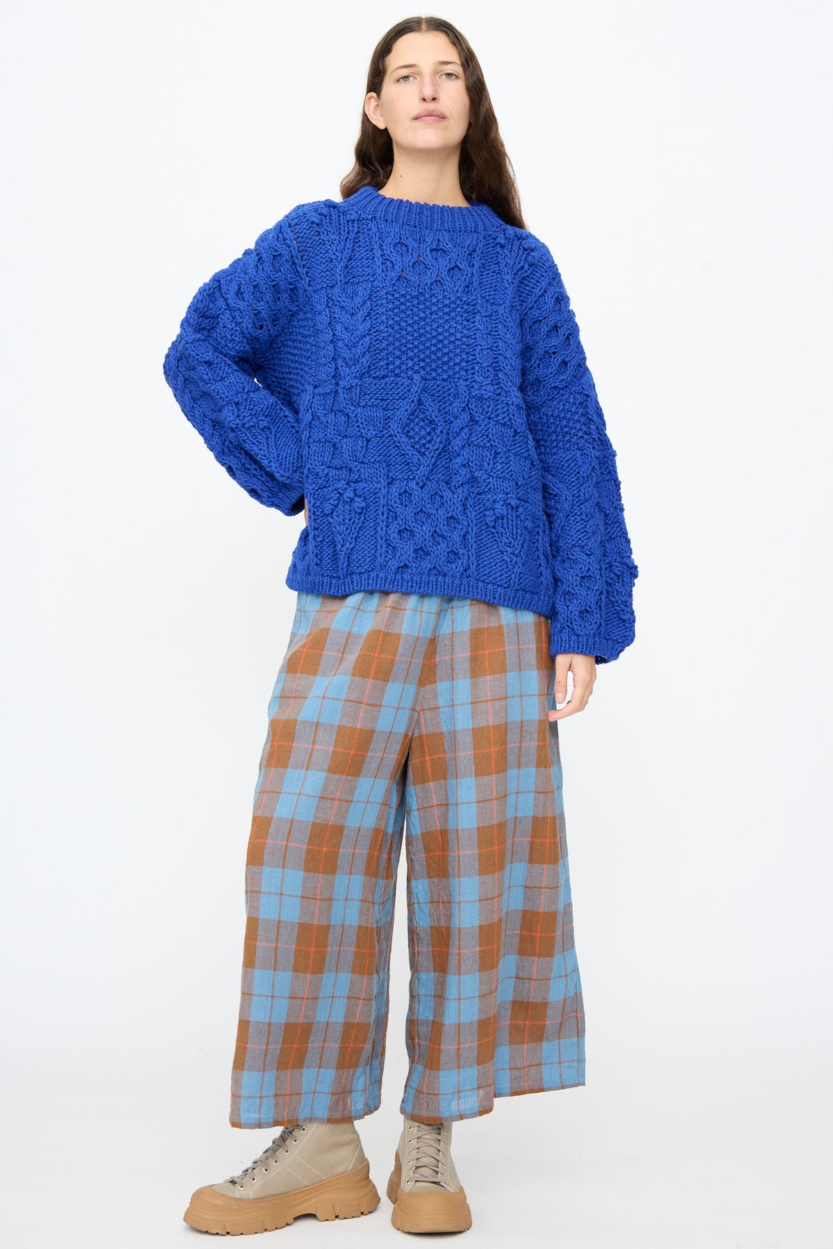 A person wears an Ichi Antiquités Wool Hand Knit Pullover in Blue, an oversized cable-knit sweater, paired with wide-leg blue and brown plaid trousers and tan lace-up boots.