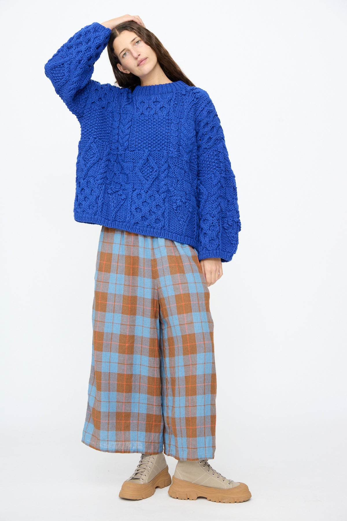 Person wearing an oversized blue Wool Hand Knit Pullover in Blue, plaid wide-leg trousers, and tan lug-sole sneakers.