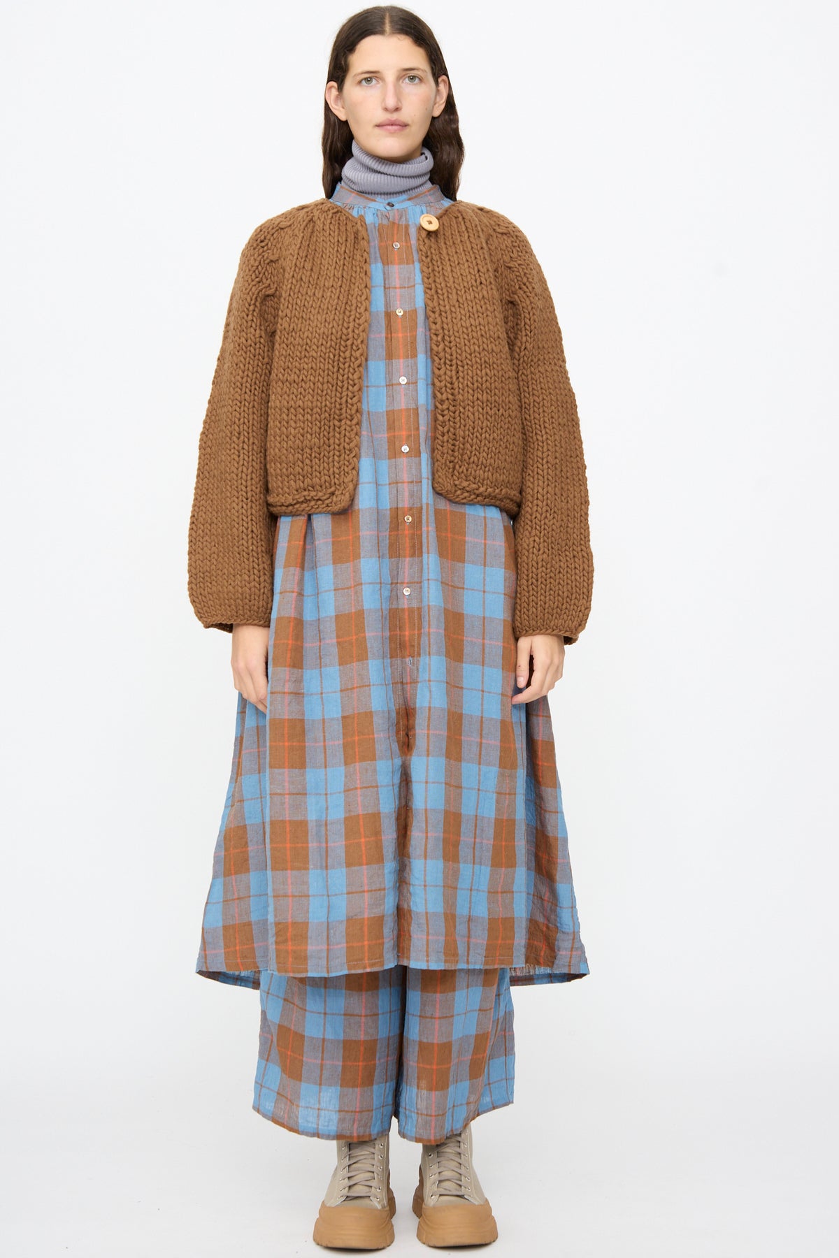 A person wearing an Ichi Antiquités Wool Hand Knit Pullover in brown over a blue and brown plaid button-front dress and matching wide-leg trousers.