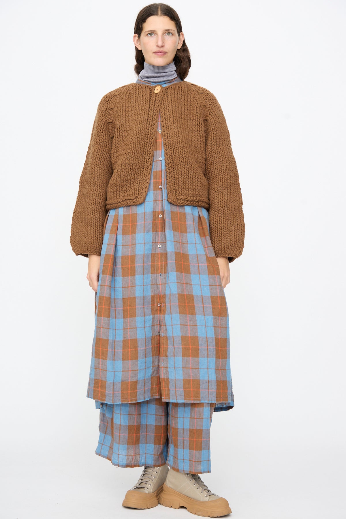 A person wearing an Ichi Antiquités Wool Hand Knit Pullover in Brown over a blue and brown plaid button-down dress and matching pants, with beige lace-up boots.