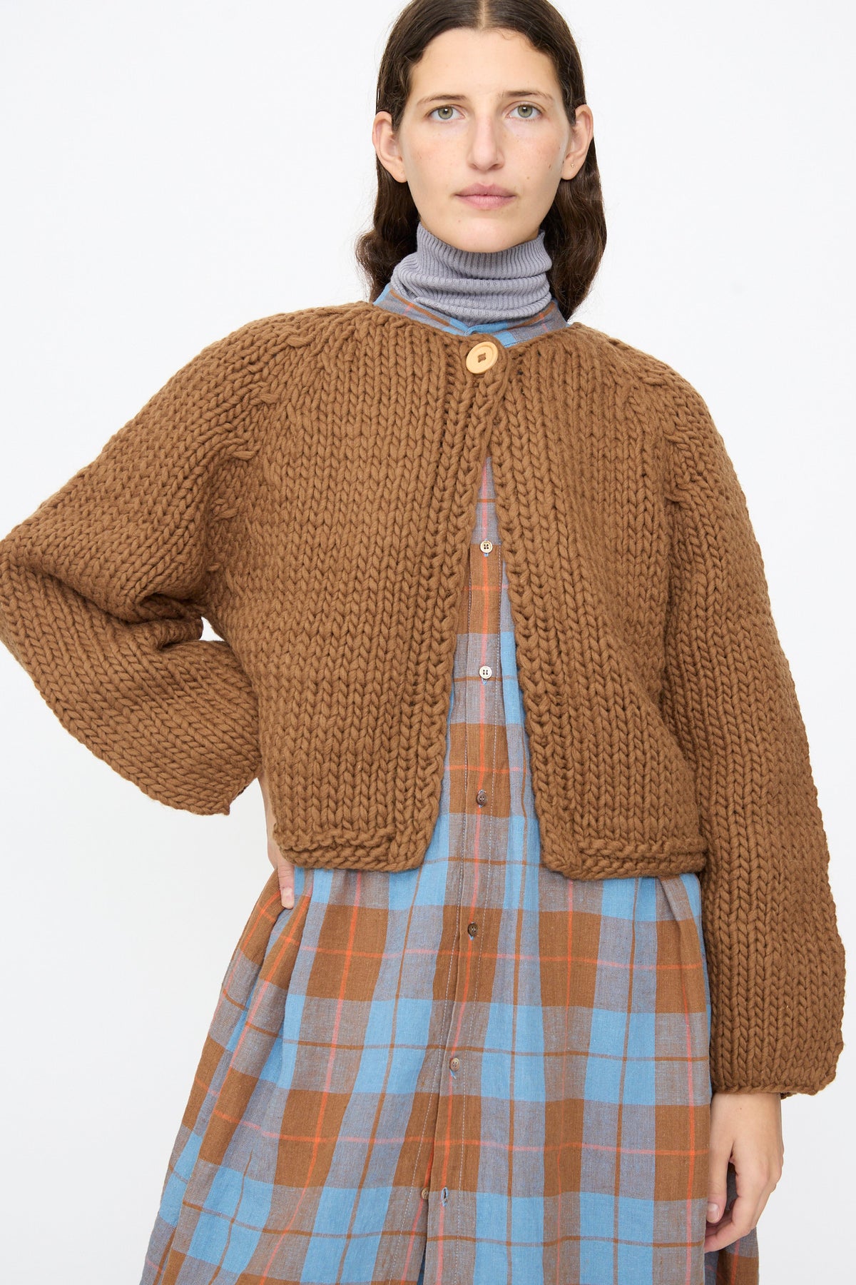 A person wears a brown Ichi Antiquités Wool Hand Knit Pullover with a large wooden button, layered over a blue and brown plaid dress and a grey turtleneck.