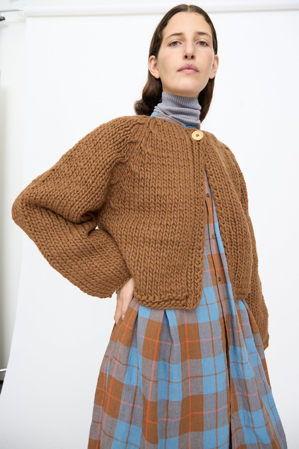 A person wearing a brown Ichi Antiquités Wool Hand Knit Pullover, a cropped, hand-knit cardigan buttoned at the neck over a grey turtleneck and a blue and brown plaid dress.