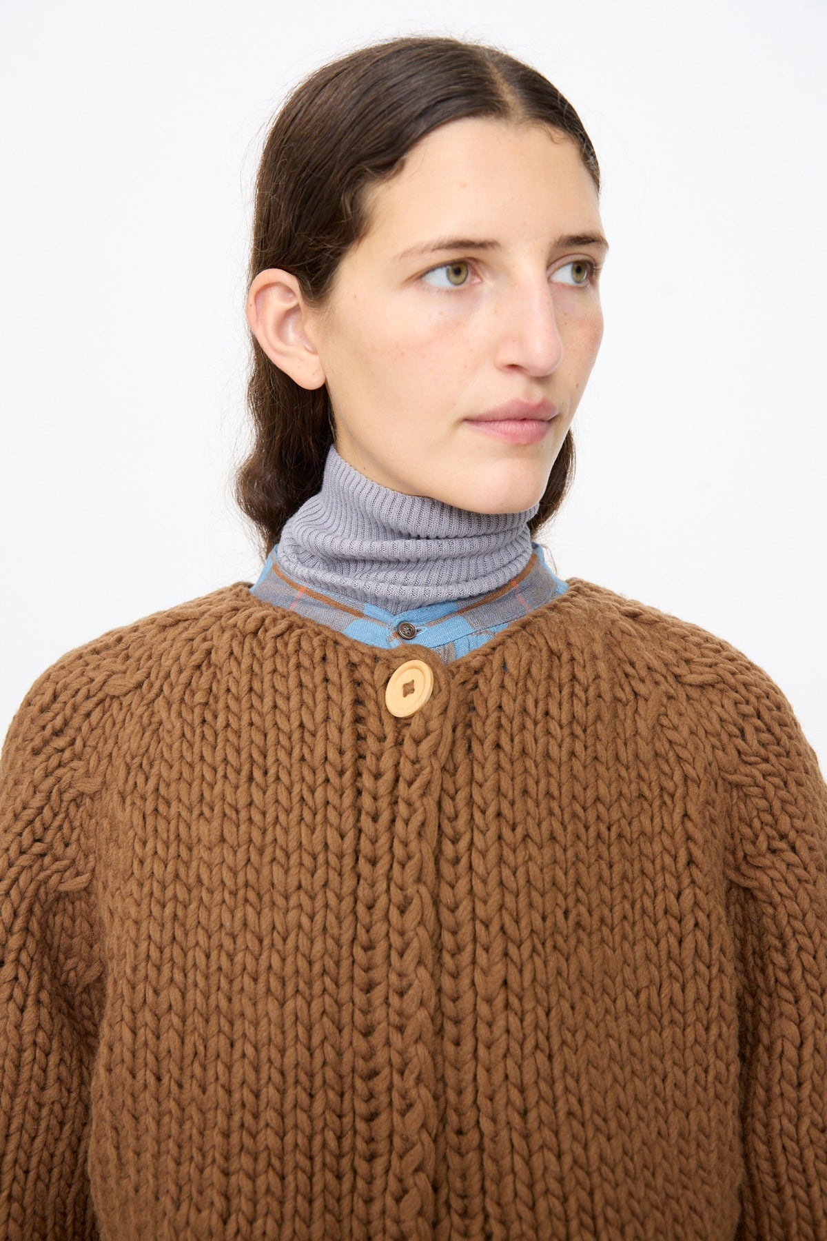A person wearing an Ichi Antiquités Wool Hand Knit Pullover in Brown, featuring a single wooden button, over a grey turtleneck and a blue plaid shirt.
