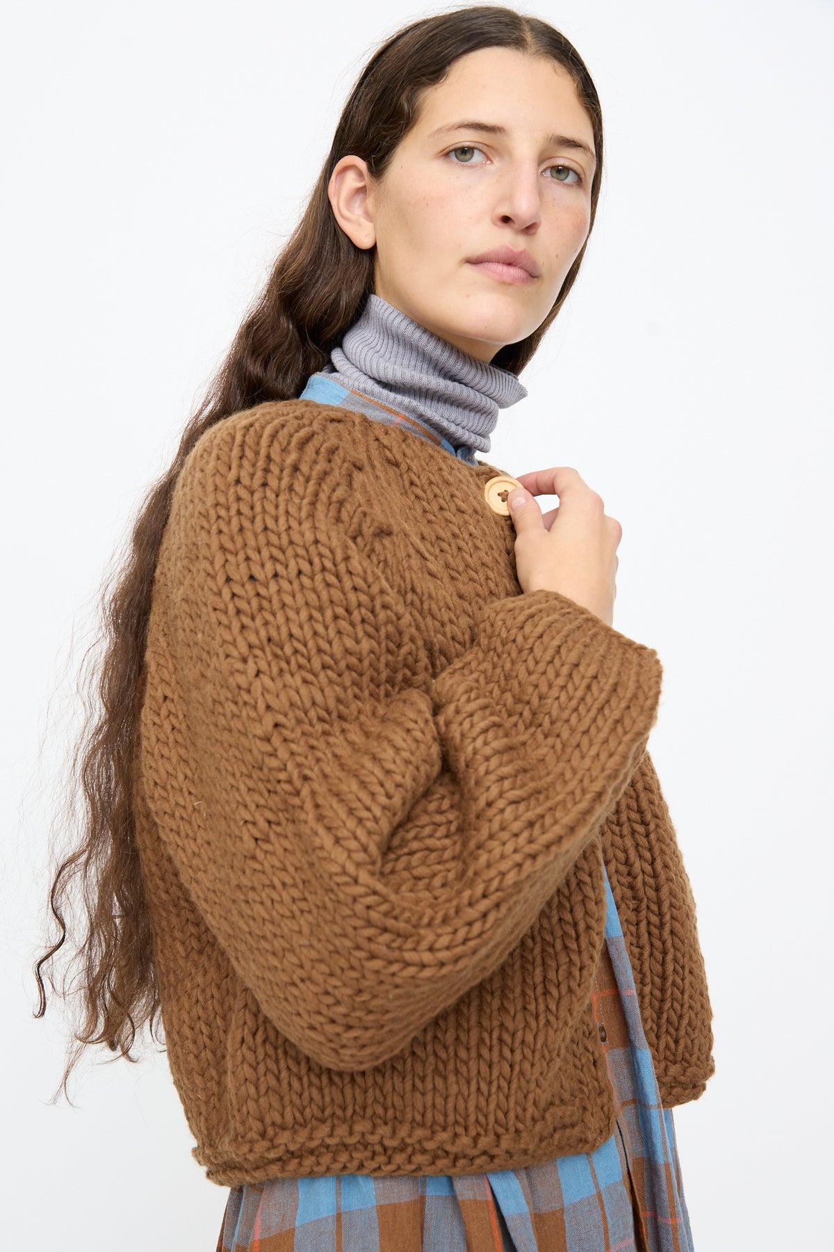 A person wears an Ichi Antiquités Wool Hand Knit Pullover in Brown, a cropped and chunky hand-knit sweater, layered over a blue plaid shirt and a grey turtleneck.
