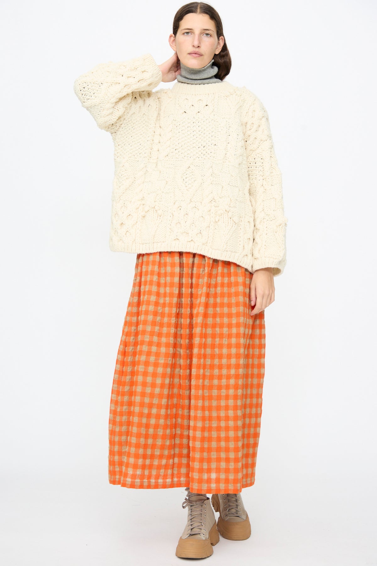 A model wears an Ichi Antiquités Wool Hand Knit Pullover in White over a grey mock-neck top and an orange and beige gingham skirt with tan lace-up boots.