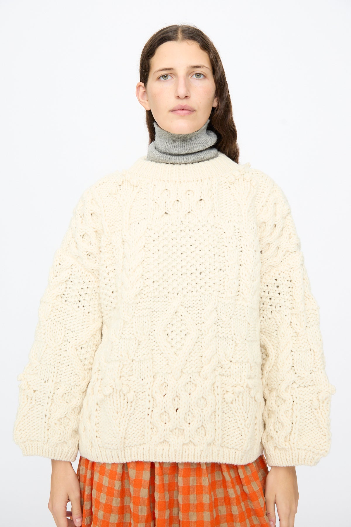 A person wearing an oversized Ichi Antiquités Wool Hand Knit Pullover in White over a gray turtleneck and an orange-and-brown checkered skirt.
