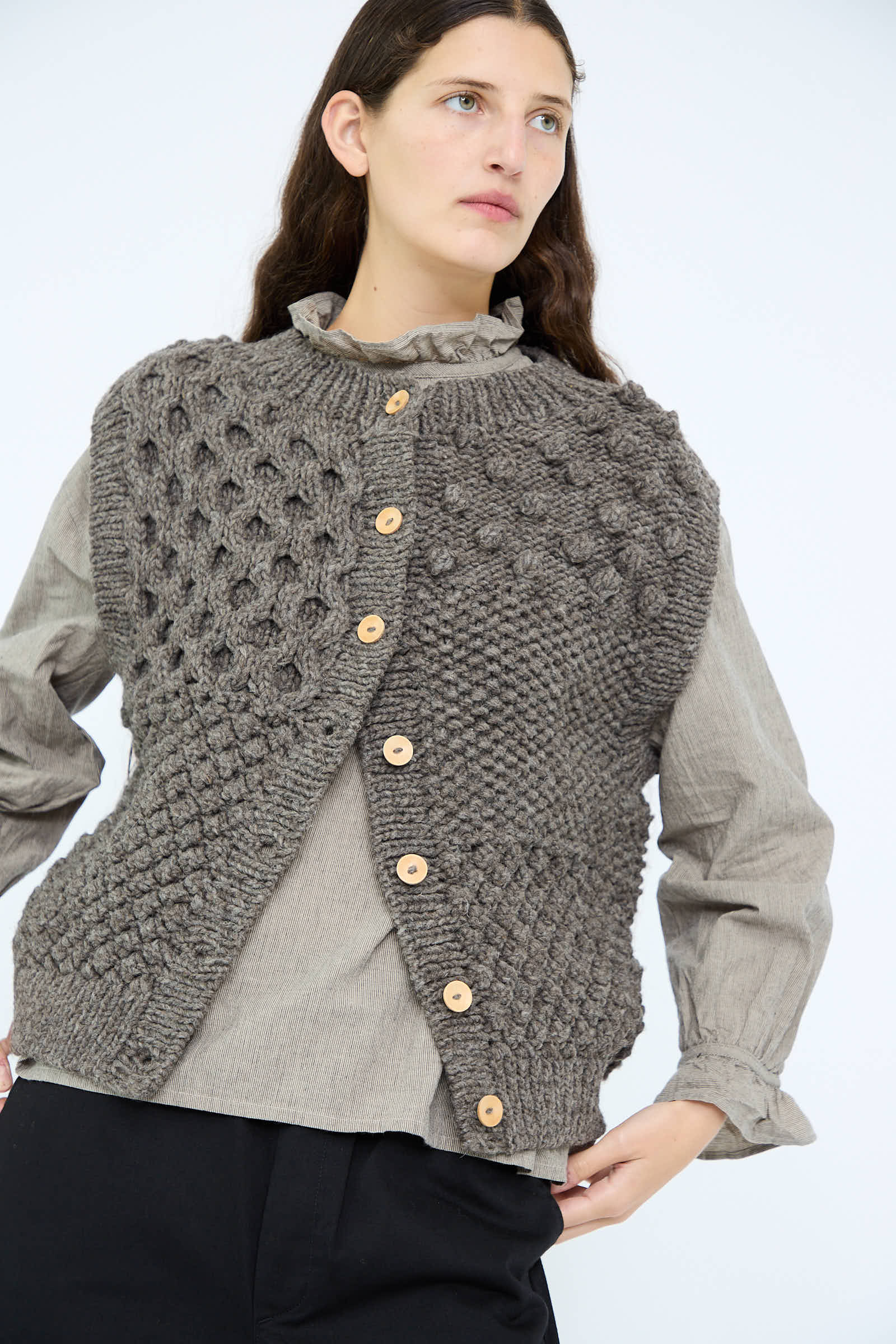 A person wears the chic Hand Knit Wool Vest in Mocha by Ichi Antiquités, featuring wooden buttons, over a gray blouse and black pants, set against a plain background.