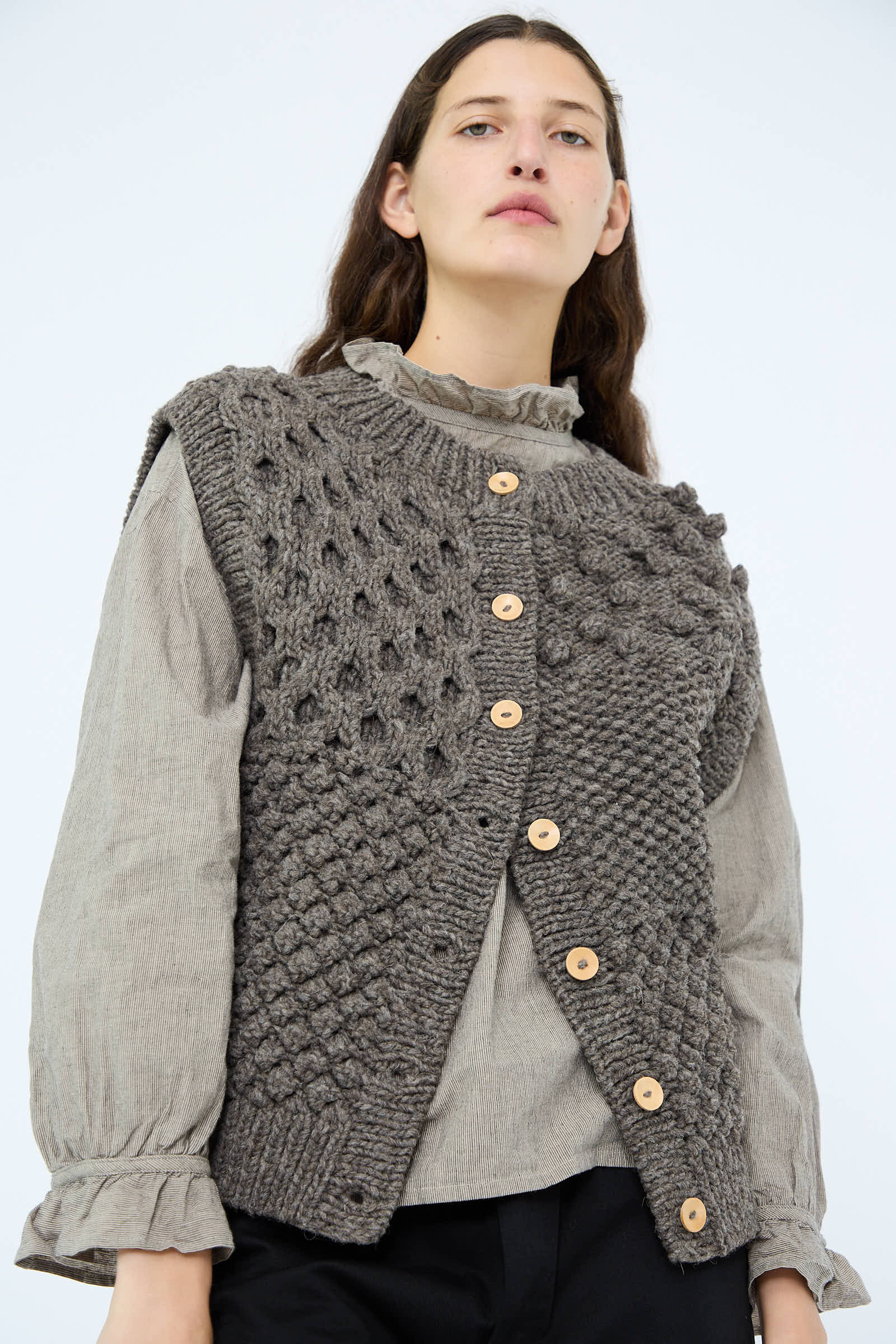 An individual wears a Hand Knit Wool Vest in Mocha by Ichi Antiquités, distinguished by its textured design and wooden buttons, layered over a light gray shirt.