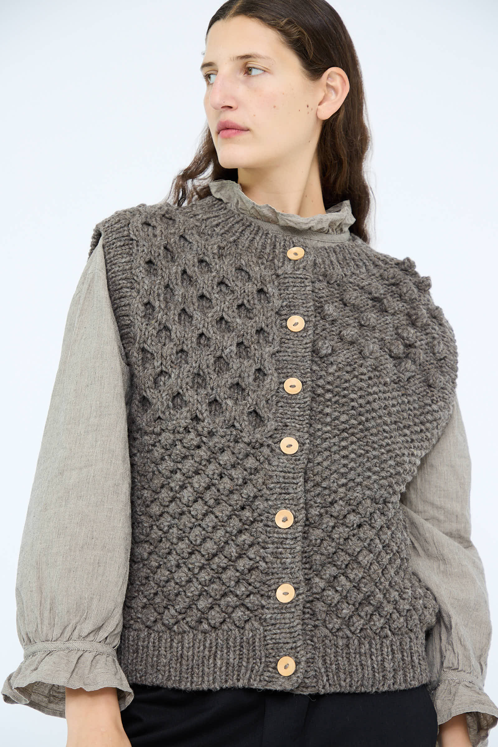 A person confidently wears the Hand Knit Wool Vest in Mocha by Ichi Antiquités over a ruffled, long-sleeve blouse.