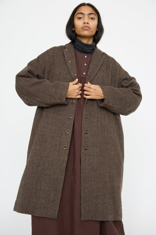 A person is wearing the Wool Herringbone Coat in Brown by Ichi Antiquités over a brown dress, standing against a plain background.