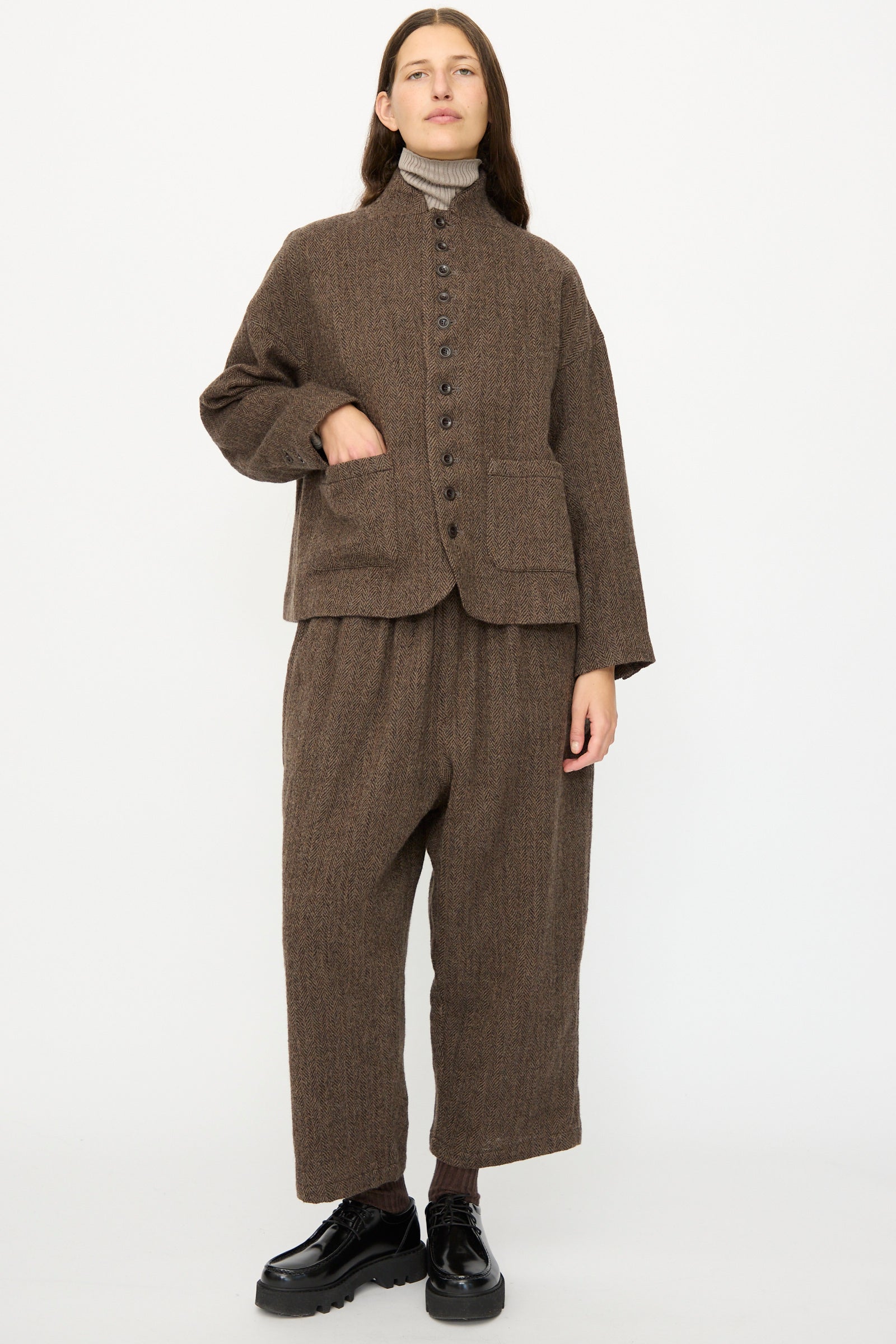 A person stands against a white background wearing the Ichi Antiquités Wool Herringbone Jacket in Brown, paired with matching wide-leg trousers, a beige turtleneck, and black shoes.