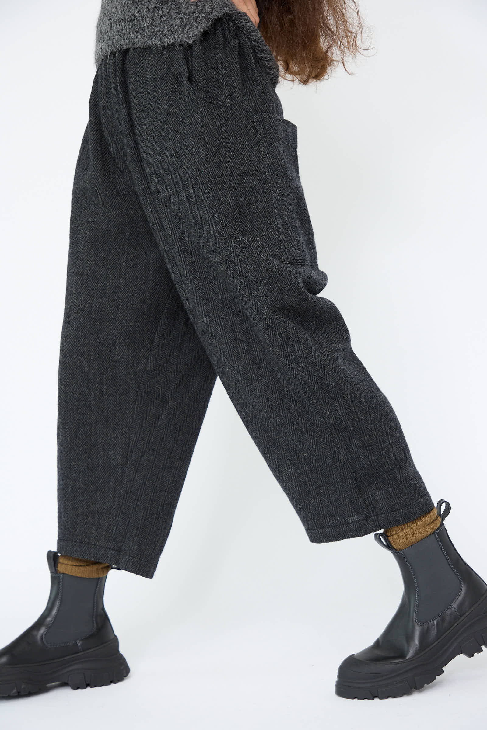 A person walks against a white background, wearing the Ichi Antiquités Wool Herringbone Pant in Black with an elasticated waist, mustard socks, and black boots.