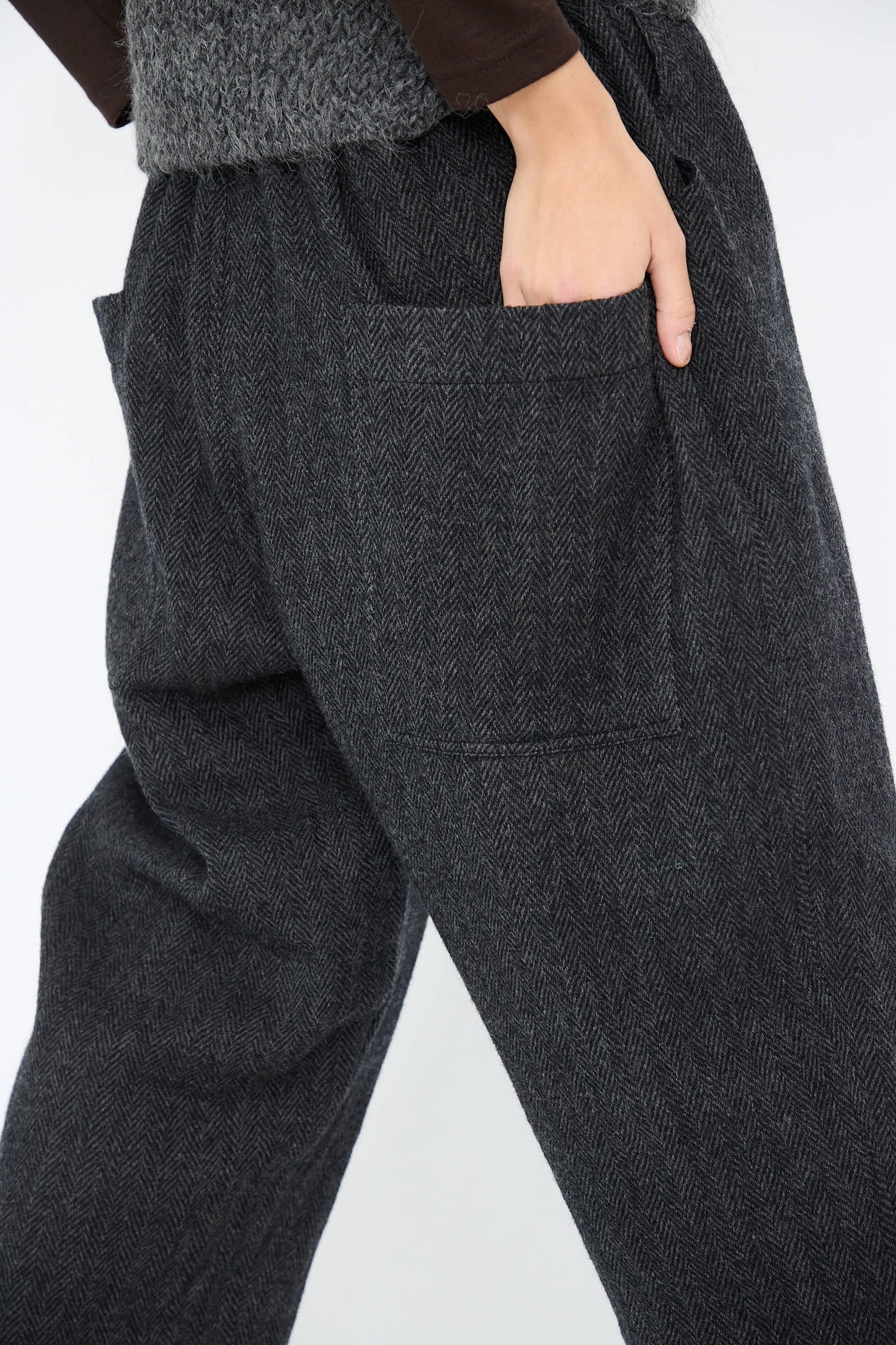 A person wearing the Wool Herringbone Pant in Black by Ichi Antiquités, featuring a charcoal herringbone pattern and an elasticated waist, casually resting a hand in the pocket against a plain background.