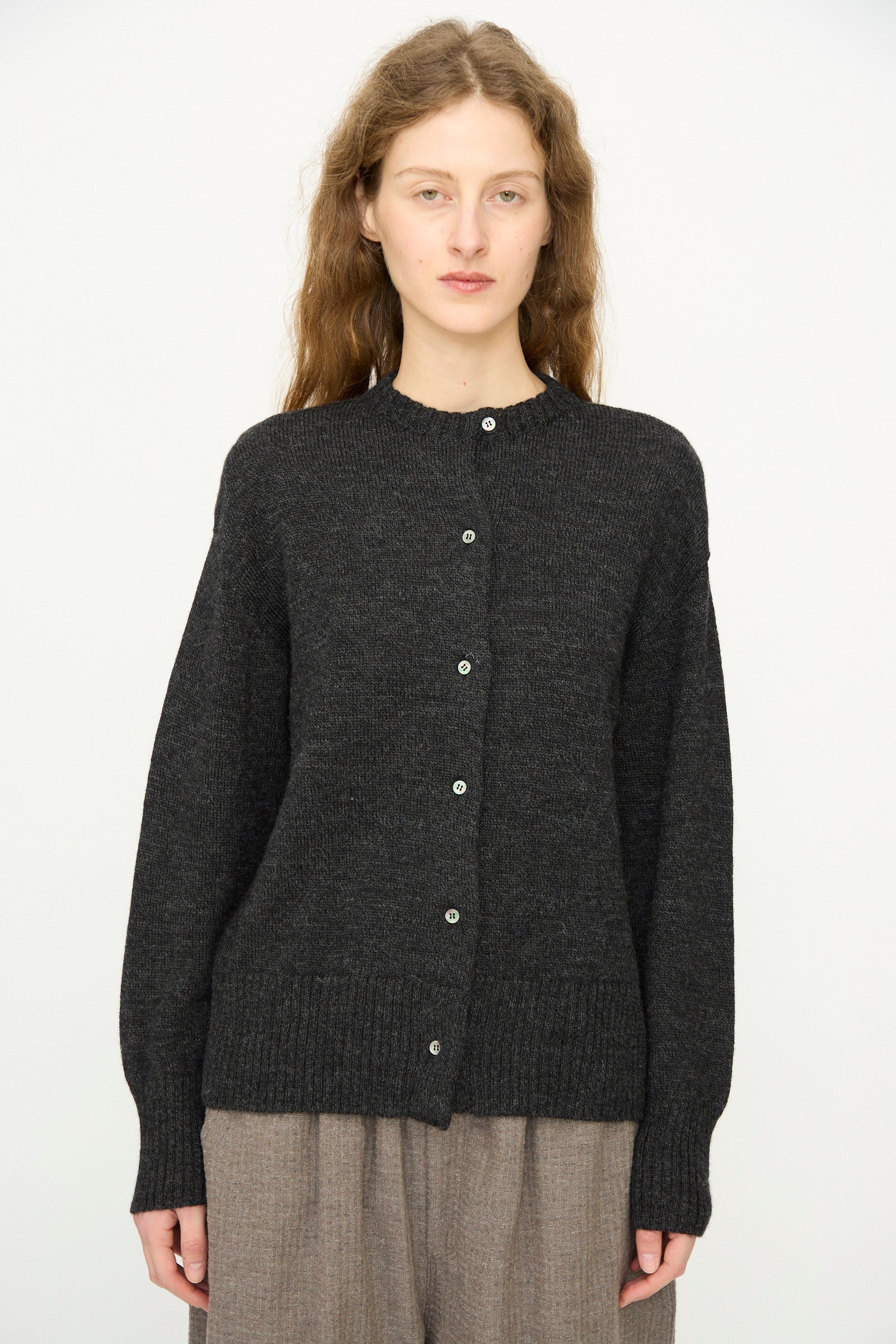 A woman with long wavy hair wears the Ichi Antiquités Wool Knit Cardigan in Black, paired with loose brown pants, standing against a plain white background.