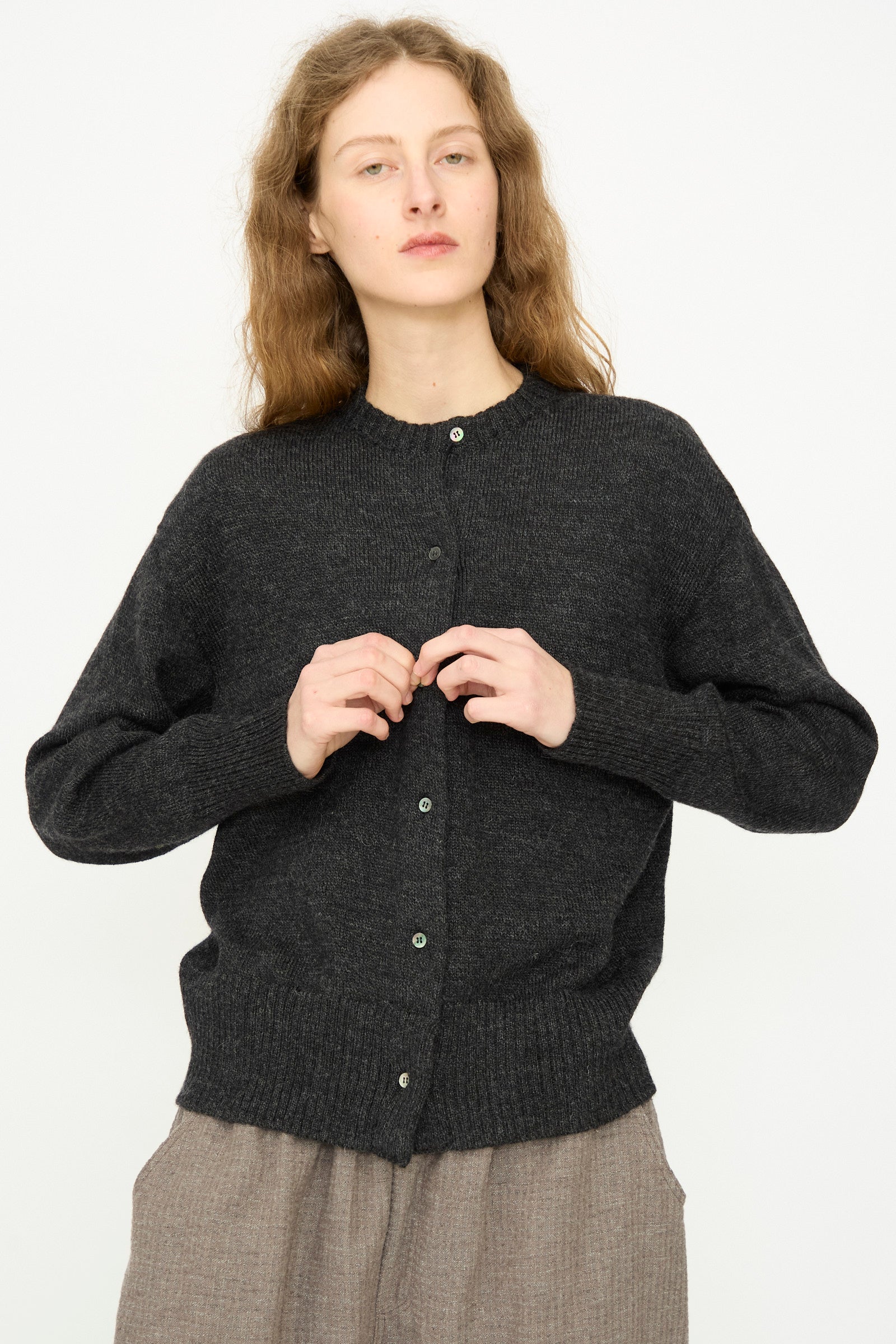 A person with wavy light brown hair wears the Ichi Antiquités Wool Knit Cardigan in Black and light brown pants, standing against a plain light background.