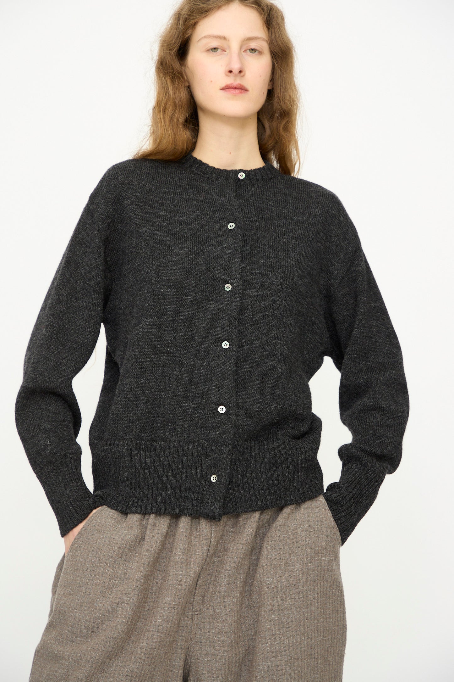 A person stands against a plain background, wearing the Ichi Antiquités Wool Knit Cardigan in Black, made in Japan, paired with light brown pants and hands in pockets.