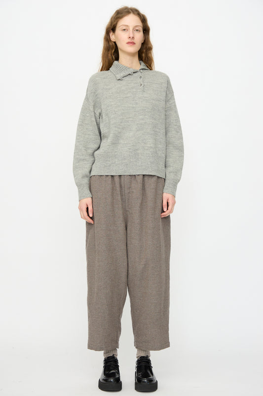 A person stands against a white background, wearing the Ichi Antiquités Wool Knit Pullover in Gray with a ribbed collar, paired with loose brown pants and black shoes.