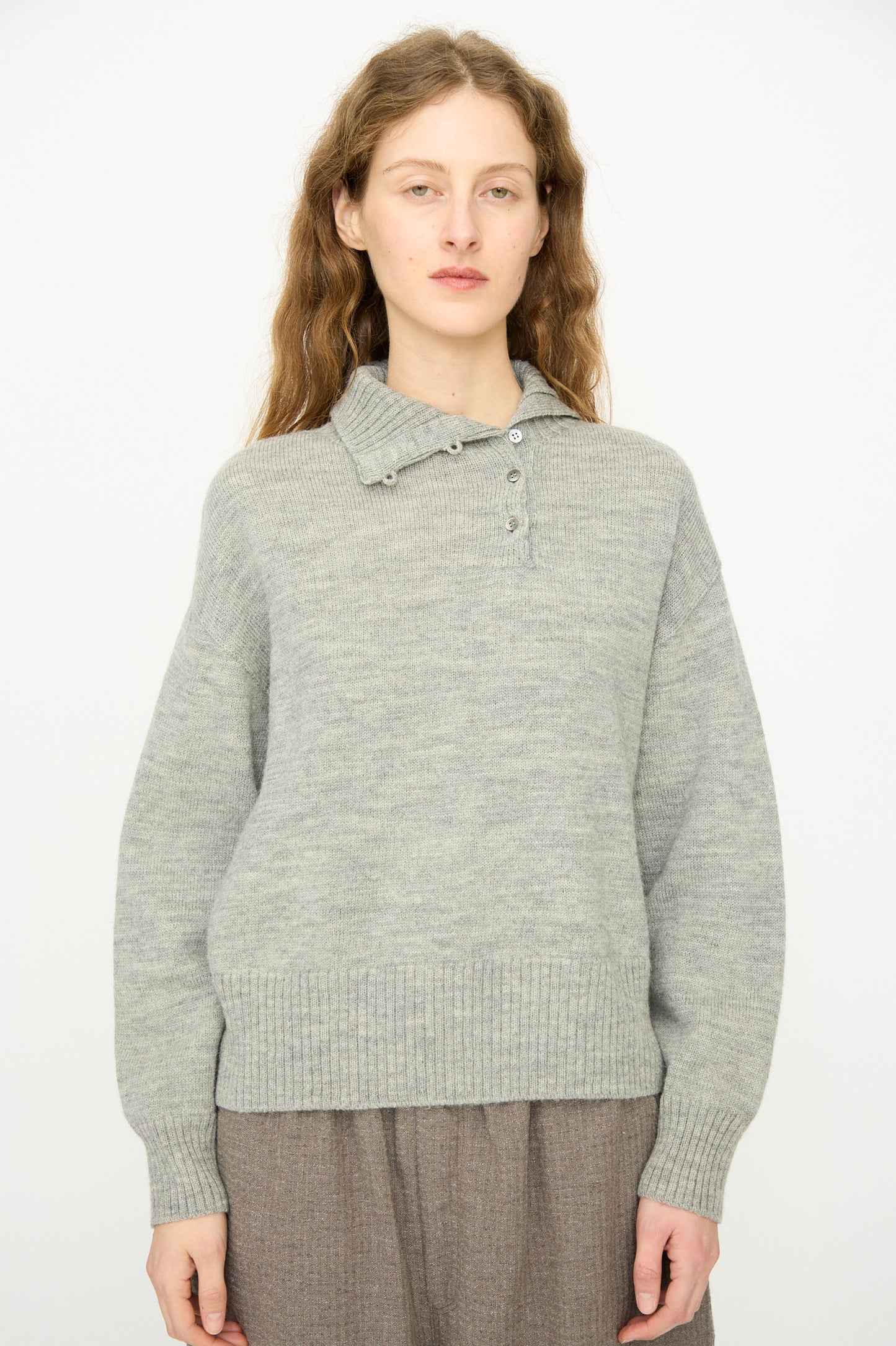 A person with long wavy hair wears the Ichi Antiquités Wool Knit Pullover in Gray, featuring button details at the ribbed collar, paired with a brown skirt, standing against a plain light background.