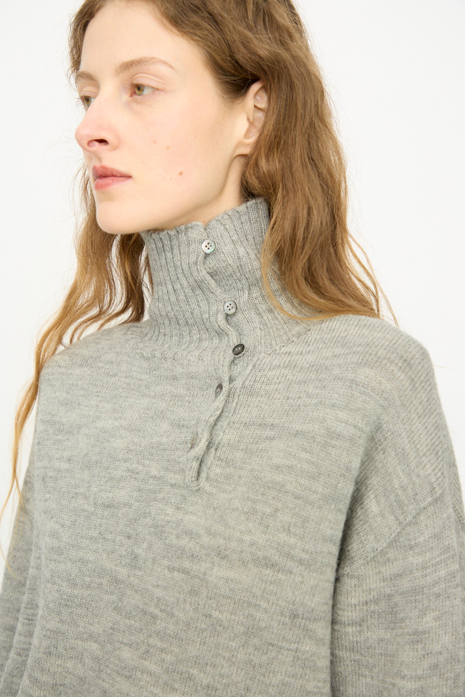 A person with long brown hair wears the Ichi Antiquités Wool Knit Pullover in Gray, featuring three visible buttons on its ribbed collar, while looking off to the side against a plain light background.