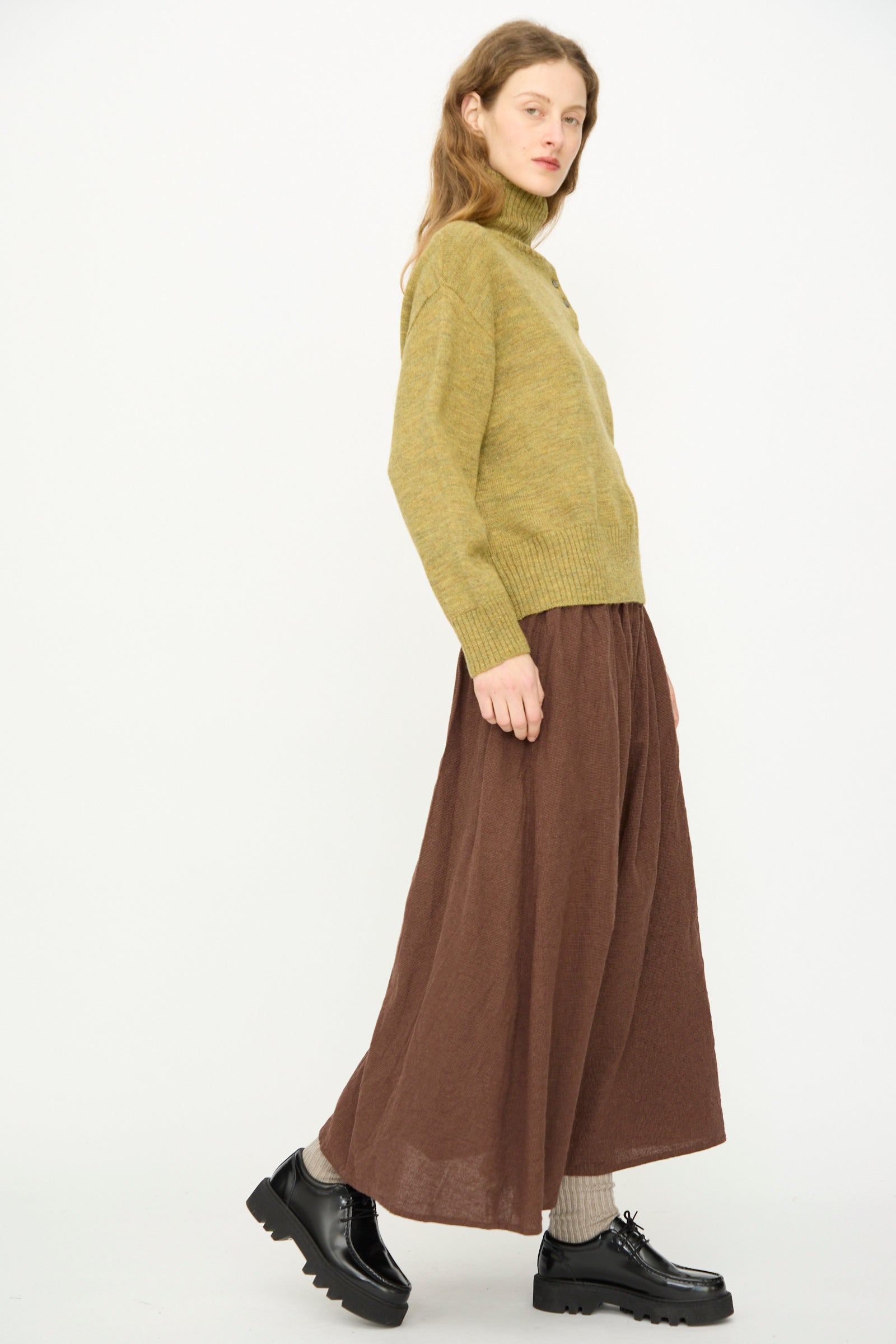 Person standing sideways against a white background, wearing the Ichi Antiquités Wool Whole Garment Knit Pullover in Mustard, a brown wide-leg skirt, beige socks, and black lace-up shoes.