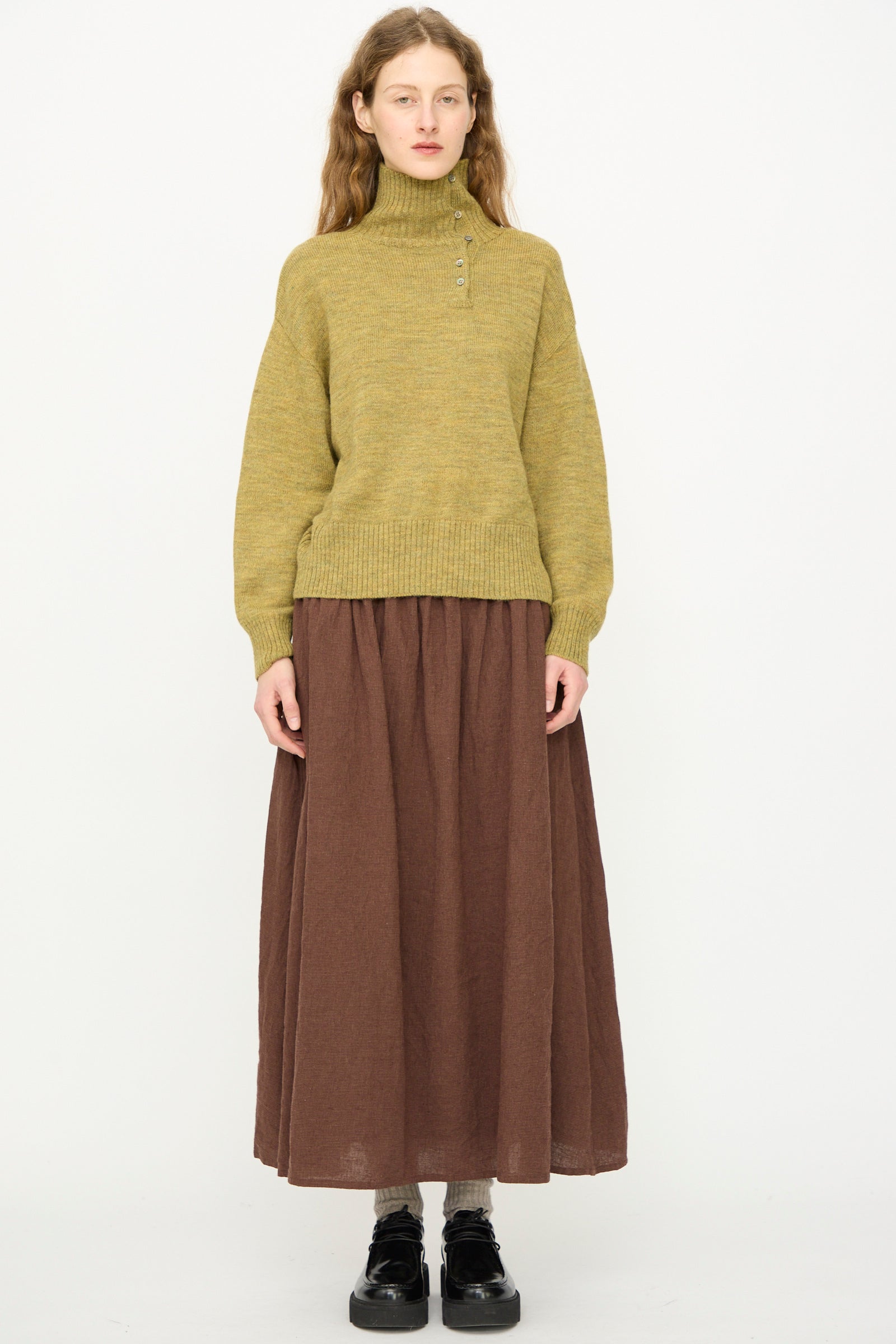 A person stands against a plain background wearing the Ichi Antiquités Wool Whole Garment Knit Pullover in Mustard, styled with a long brown skirt and black shoes. 