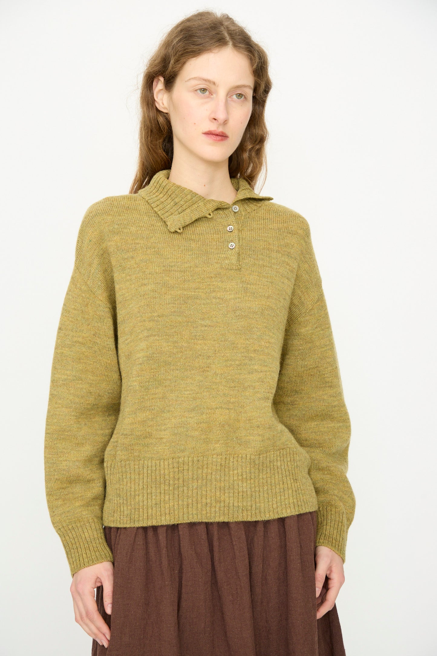 A person with long hair wears the Ichi Antiquités Wool Whole Garment Knit Pullover in Mustard paired with a brown skirt, standing against a plain light background. 
