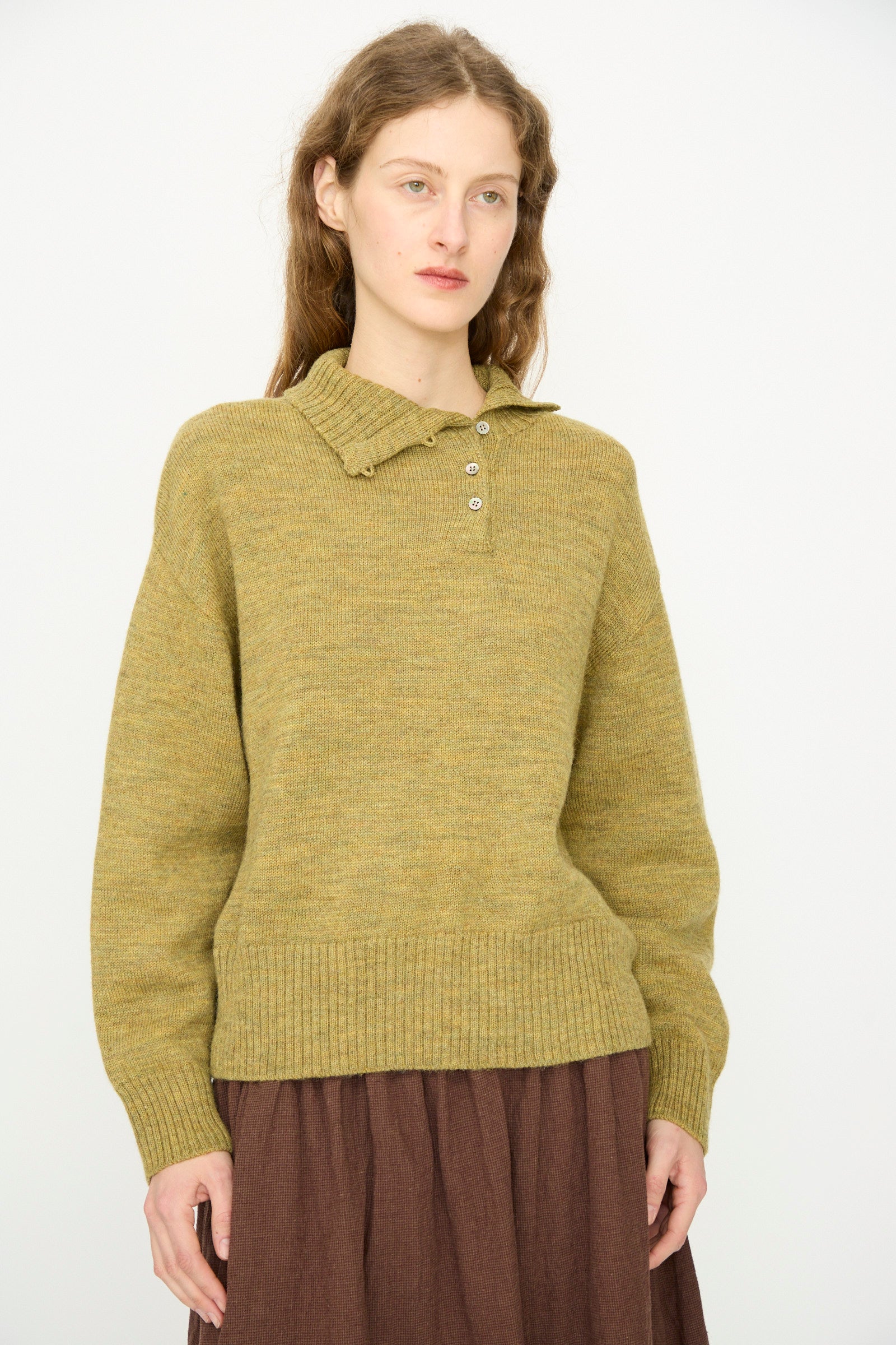 A person with long hair wears the Ichi Antiquités Wool Whole Garment Knit Pullover in Mustard paired with a brown skirt, standing against a plain light background. 