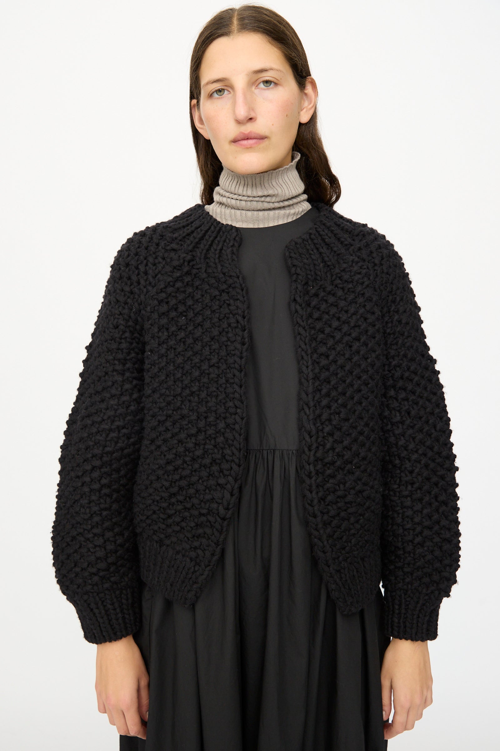 A woman stands against a plain white background wearing the Ichi Antiquités Wool Peru Hand-knit Cardigan in Black over a black dress and a light grey turtleneck.