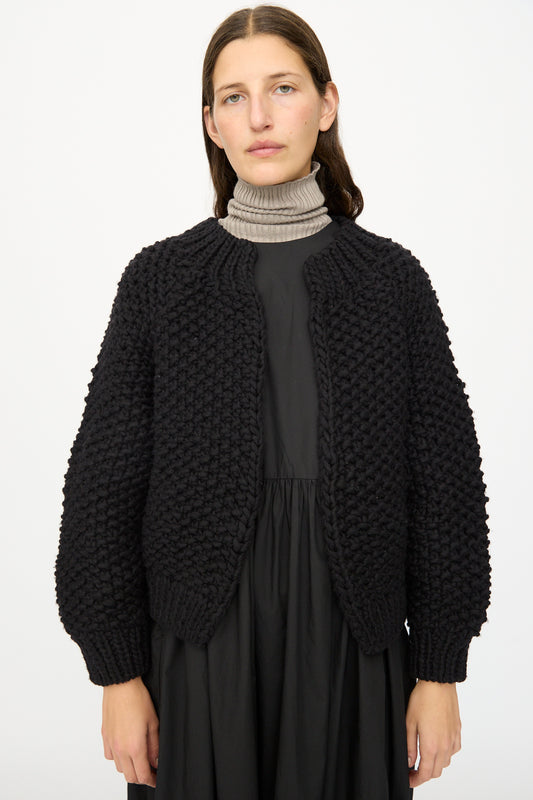 A woman stands against a plain white background wearing the Ichi Antiquités Wool Peru Hand-knit Cardigan in Black over a black dress and a light grey turtleneck.