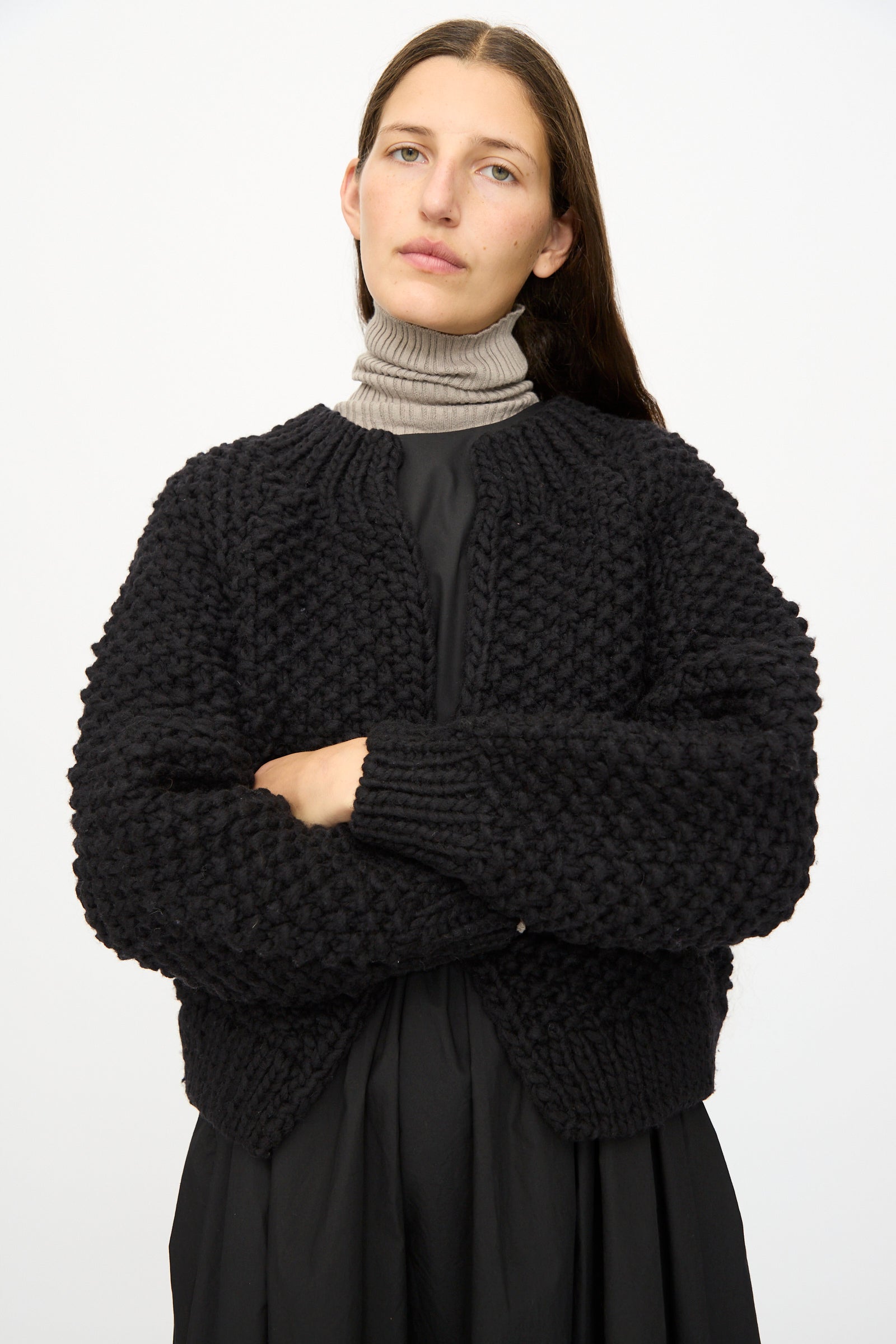 A person with long brown hair stands with arms crossed against a plain white background, wearing the Ichi Antiquités Wool Peru Hand-knit Cardigan in Black layered over a gray turtleneck and black dress.