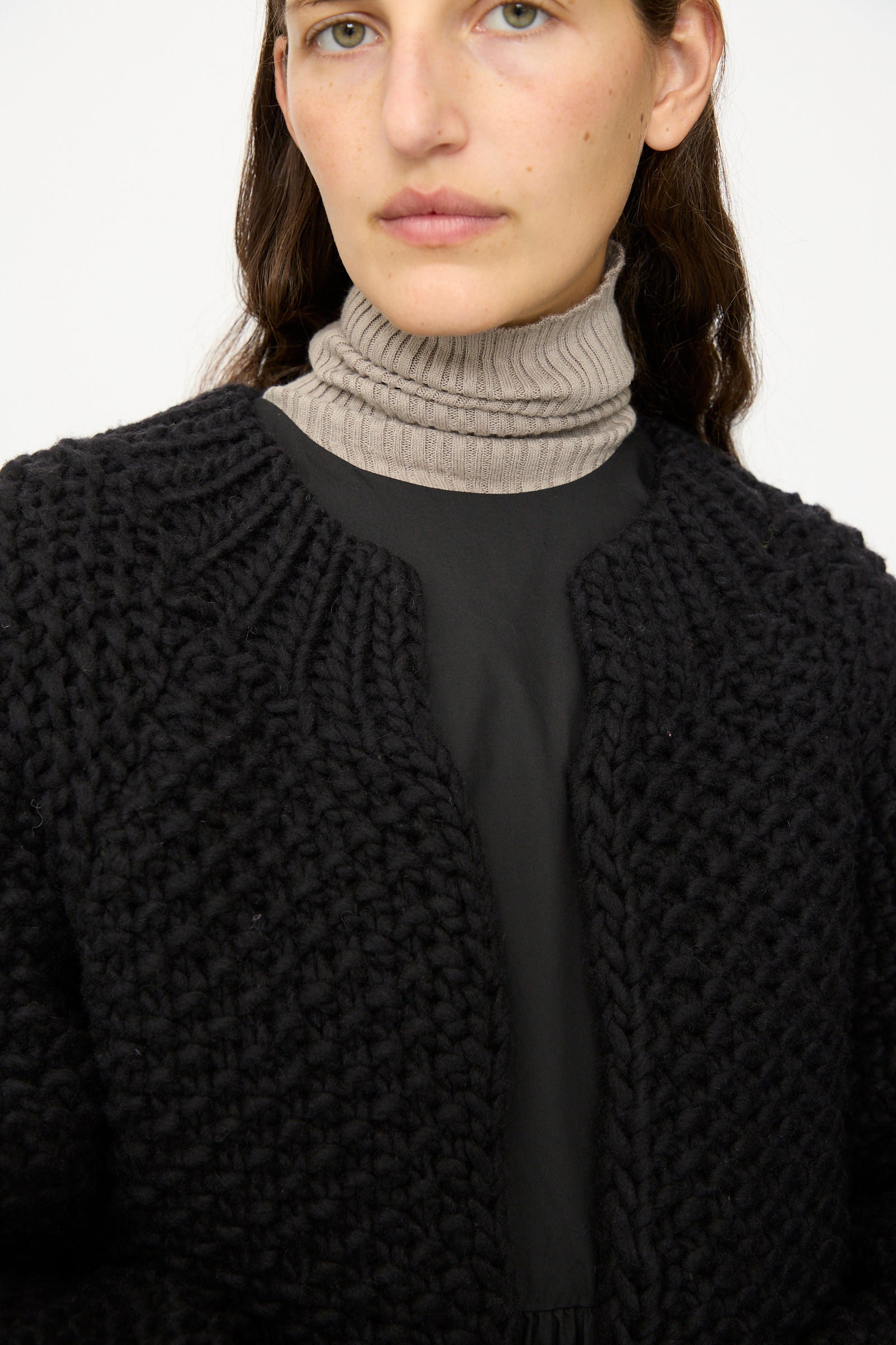 A person wears the Ichi Antiquités Wool Peru Hand-knit Cardigan in Black over a black top and beige ribbed turtleneck, facing forward with a neutral expression.