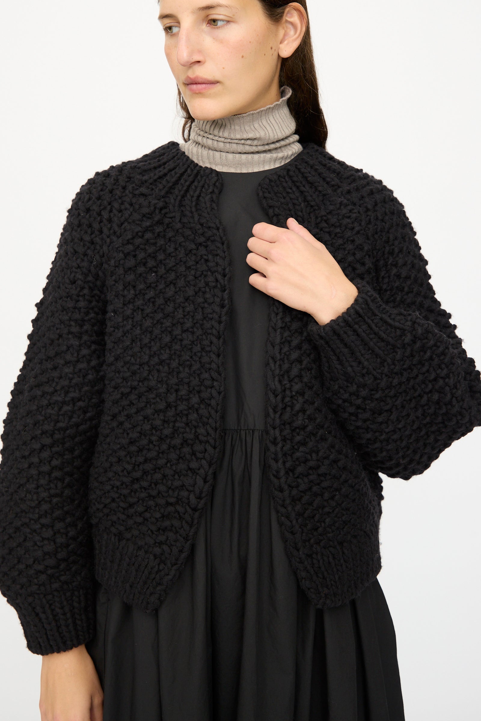 A person stands against a plain white background wearing the Ichi Antiquités Wool Peru Hand-knit Cardigan in Black over a taupe turtleneck and a black dress.