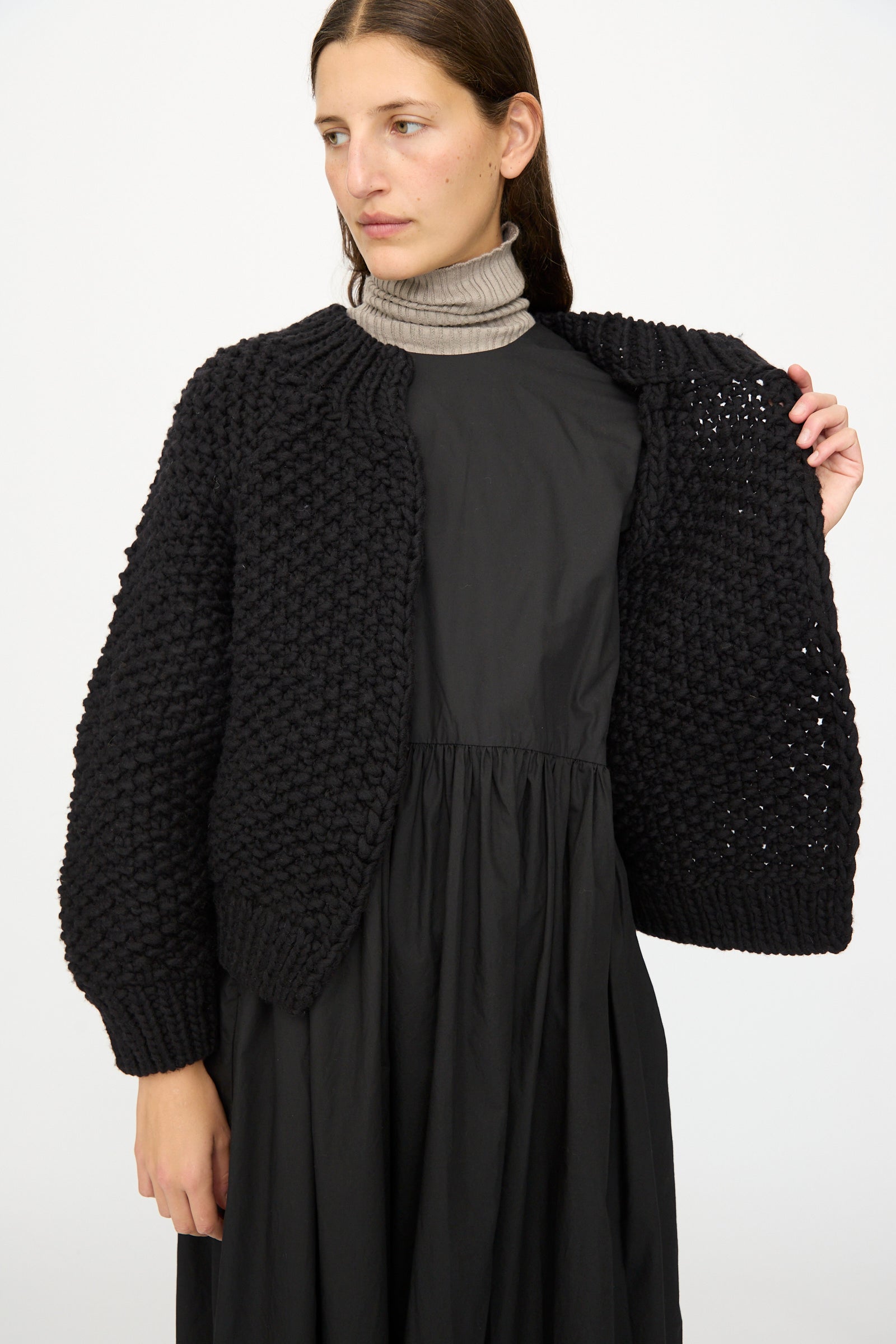 A woman in a black dress and gray turtleneck shows the interior of the Wool Peru Hand-knit Cardigan in Black by Ichi Antiquités, highlighting its hand-knit wool texture against a plain white background.