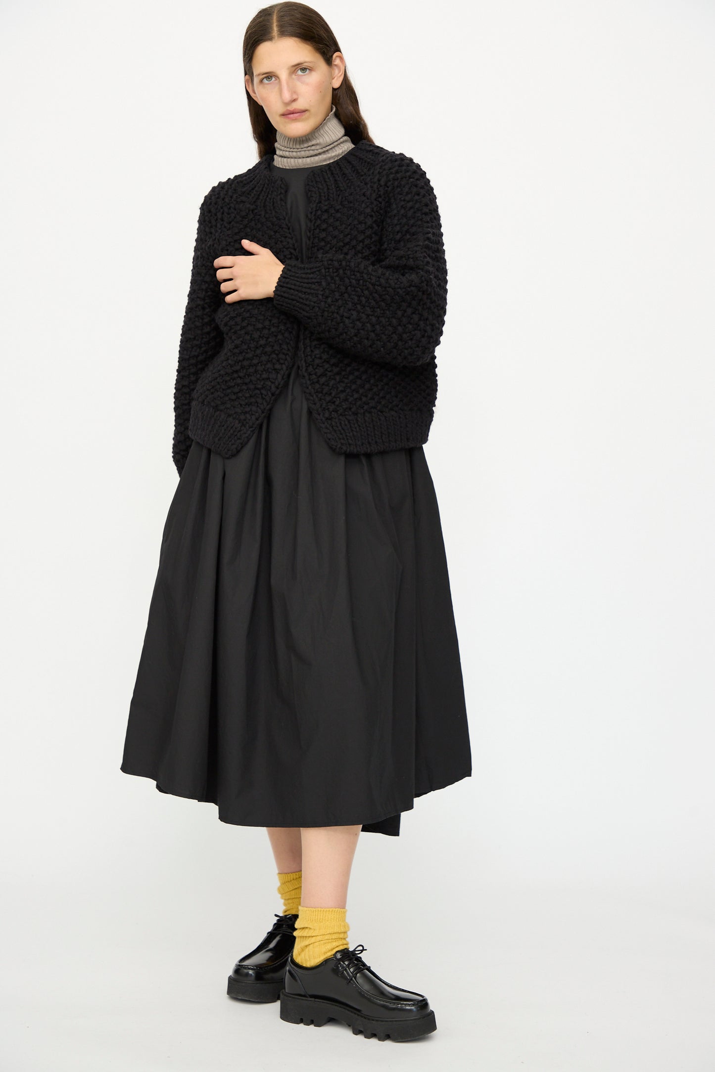 A person stands against a white background wearing the Ichi Antiquités Wool Peru Hand-knit Cardigan in Black, a black skirt, gray turtleneck, yellow socks, and black platform shoes.