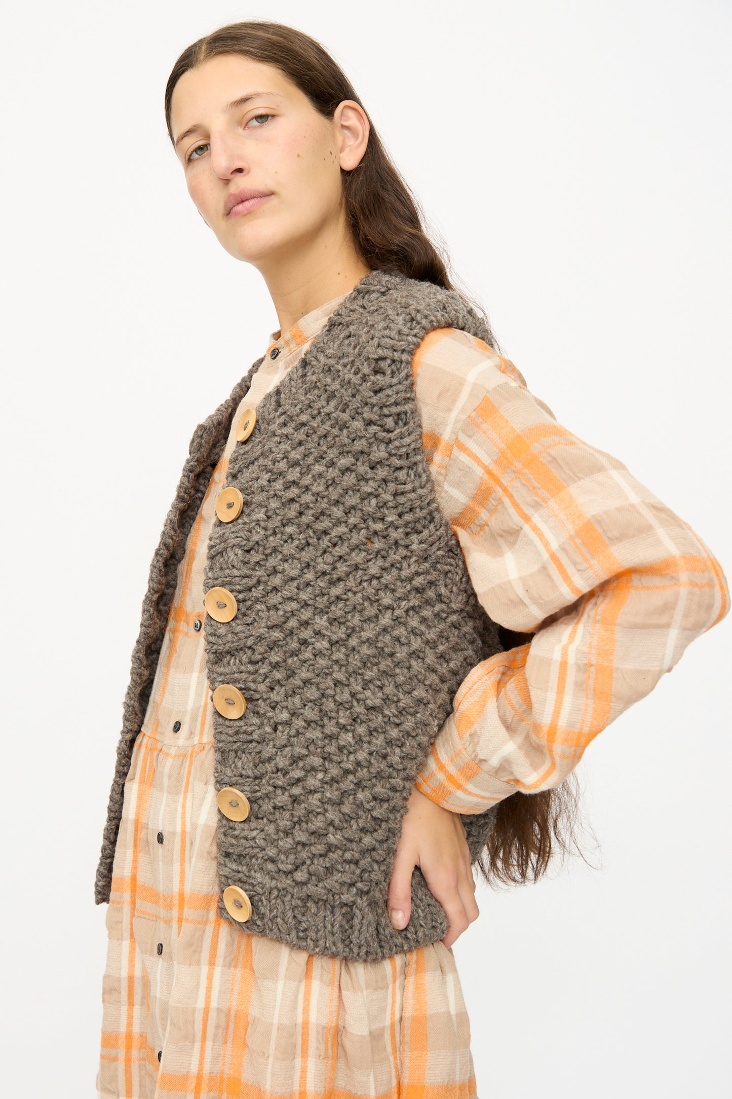 A person wearing a plaid dress with orange and beige tones and the Ichi Antiquités Wool Peru Hand-knit Vest in Gray, featuring large buttons, stands against a plain white background.