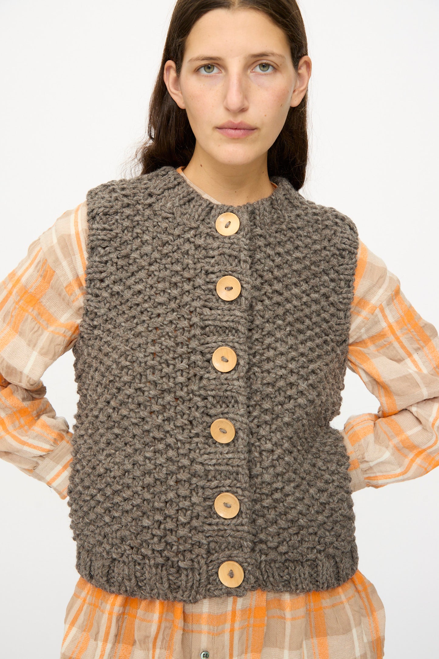 A woman wears the Ichi Antiquités Wool Peru Hand-knit Vest in Gray, featuring large wooden buttons, over a beige and orange plaid shirt against a plain white background.