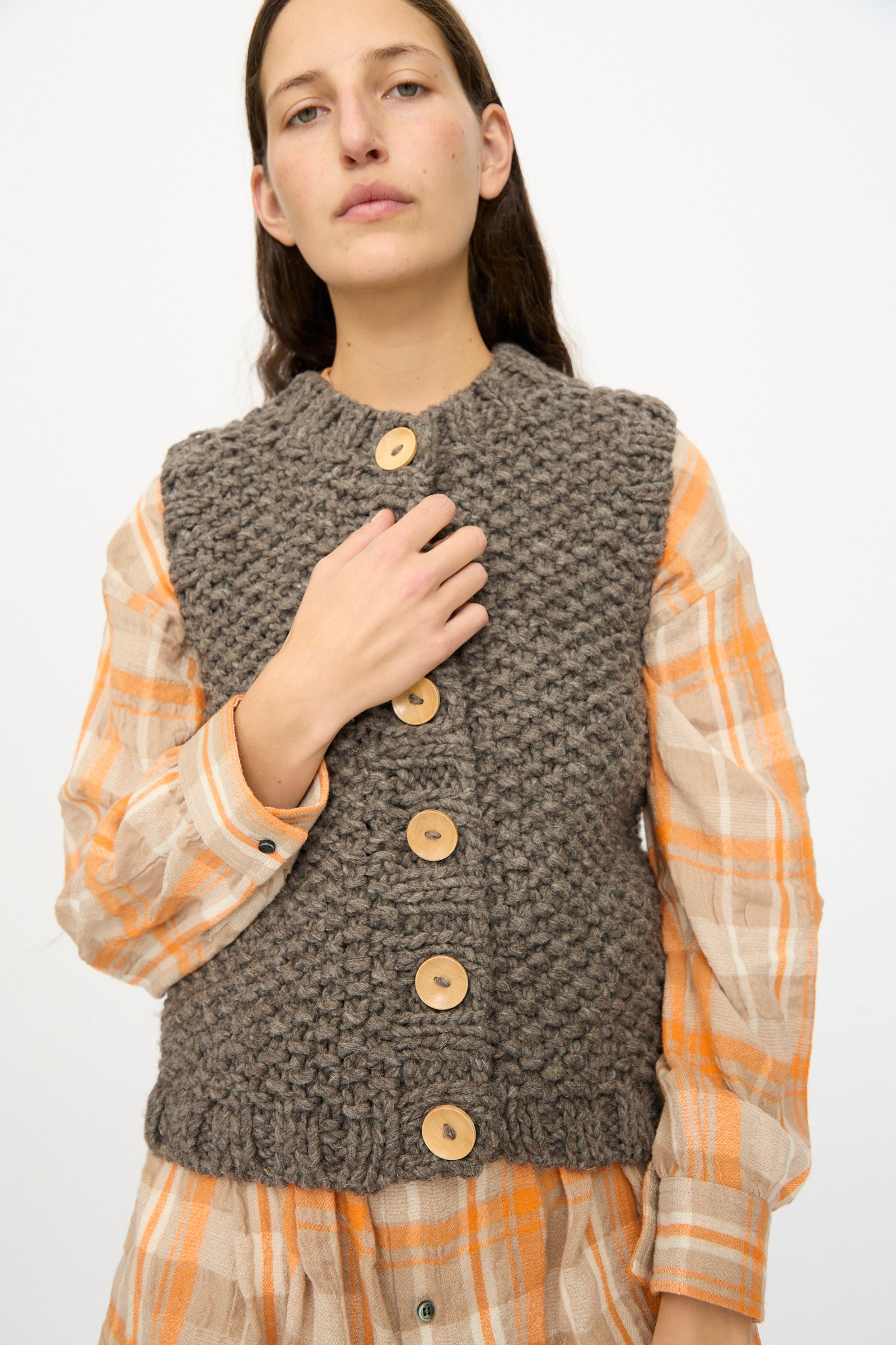 A person wearing the Ichi Antiquités Wool Peru Hand-knit Vest in Gray over an orange and beige plaid shirt, standing against a plain light background.