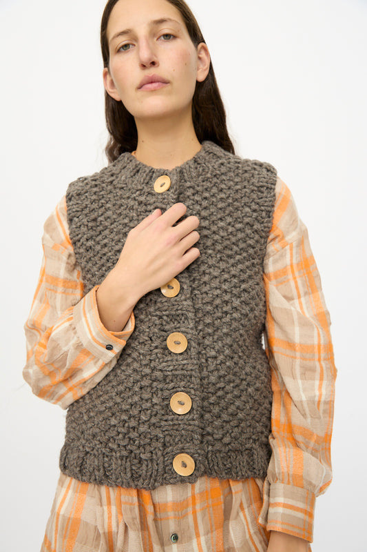 A person wearing the Ichi Antiquités Wool Peru Hand-knit Vest in Gray over an orange and beige plaid shirt, standing against a plain light background.