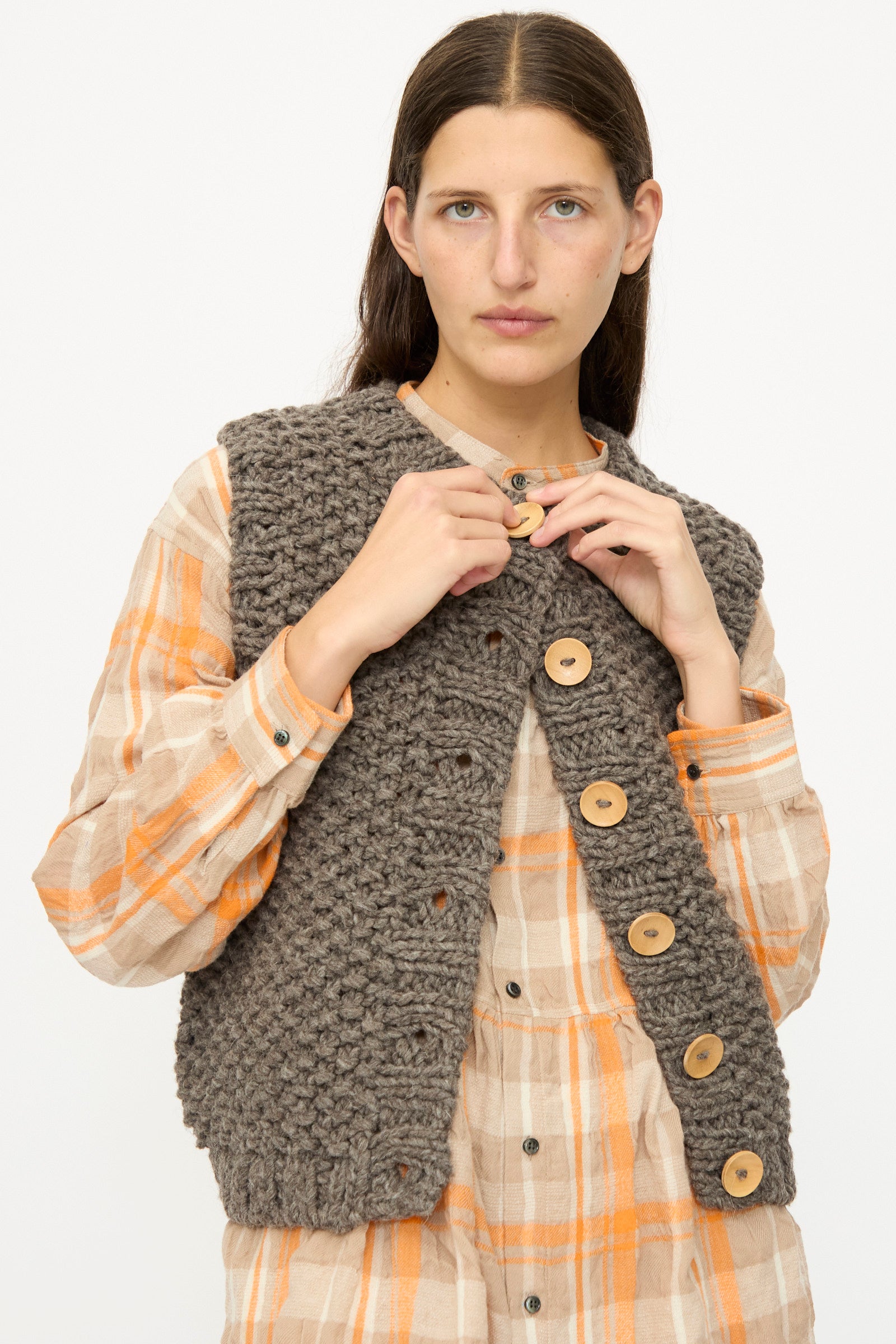 A person wears the Ichi Antiquités Wool Peru Hand-knit Vest in Gray over a beige and orange plaid button-up dress, fastening the vest’s top button.