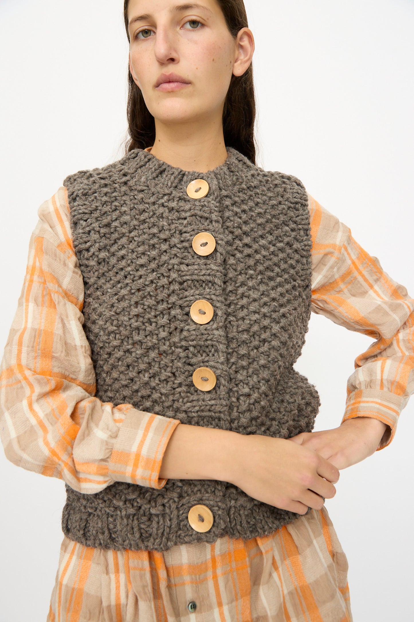A person models the Ichi Antiquités Wool Peru Hand-knit Vest in Gray, styled over a beige and orange plaid dress, posing against a plain white background.