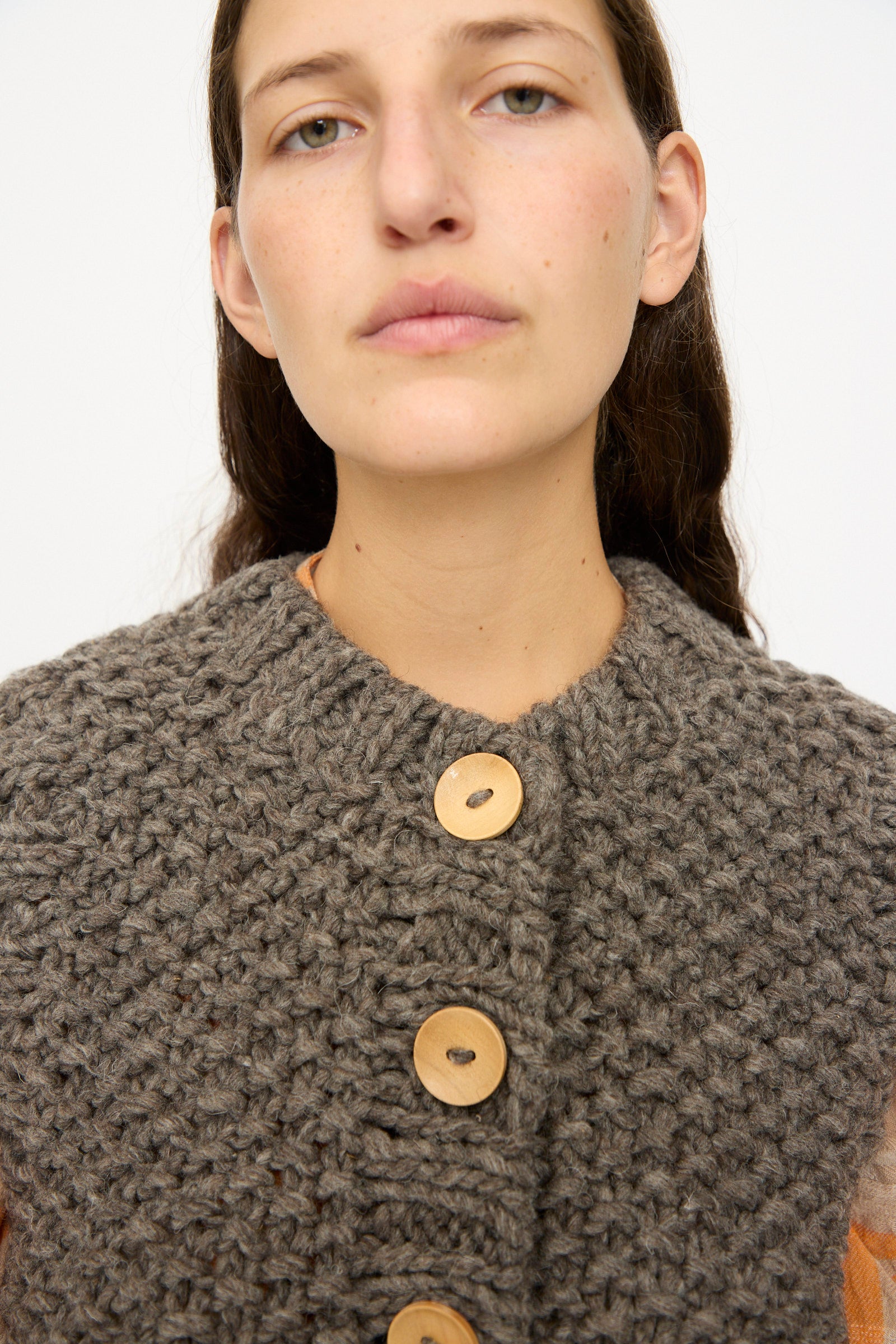 A person with long brown hair models the Ichi Antiquités Wool Peru Hand-knit Vest in Gray, featuring chunky knit and large wooden buttons, facing the camera against a plain background.
