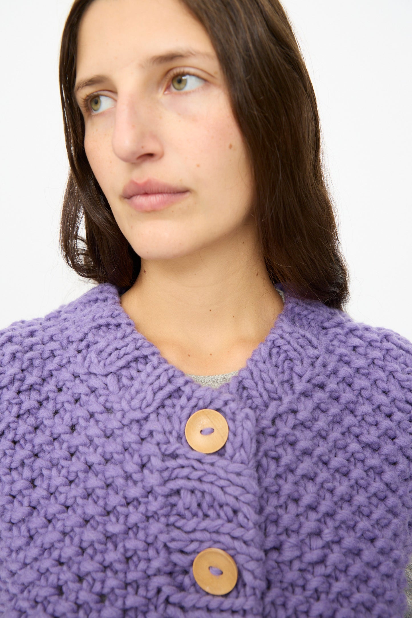 A person with long brown hair models the Ichi Antiquités Wool Peru Hand-knit Vest in Purple, featuring chunky knit and large wooden buttons, looking off to the side against a plain background.