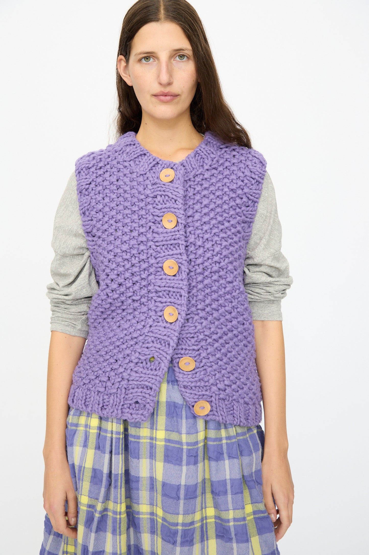 A woman wears the Ichi Antiquités Wool Peru Hand-knit Vest in Purple over a gray long-sleeve shirt and a yellow and purple plaid skirt, standing against a plain white background.