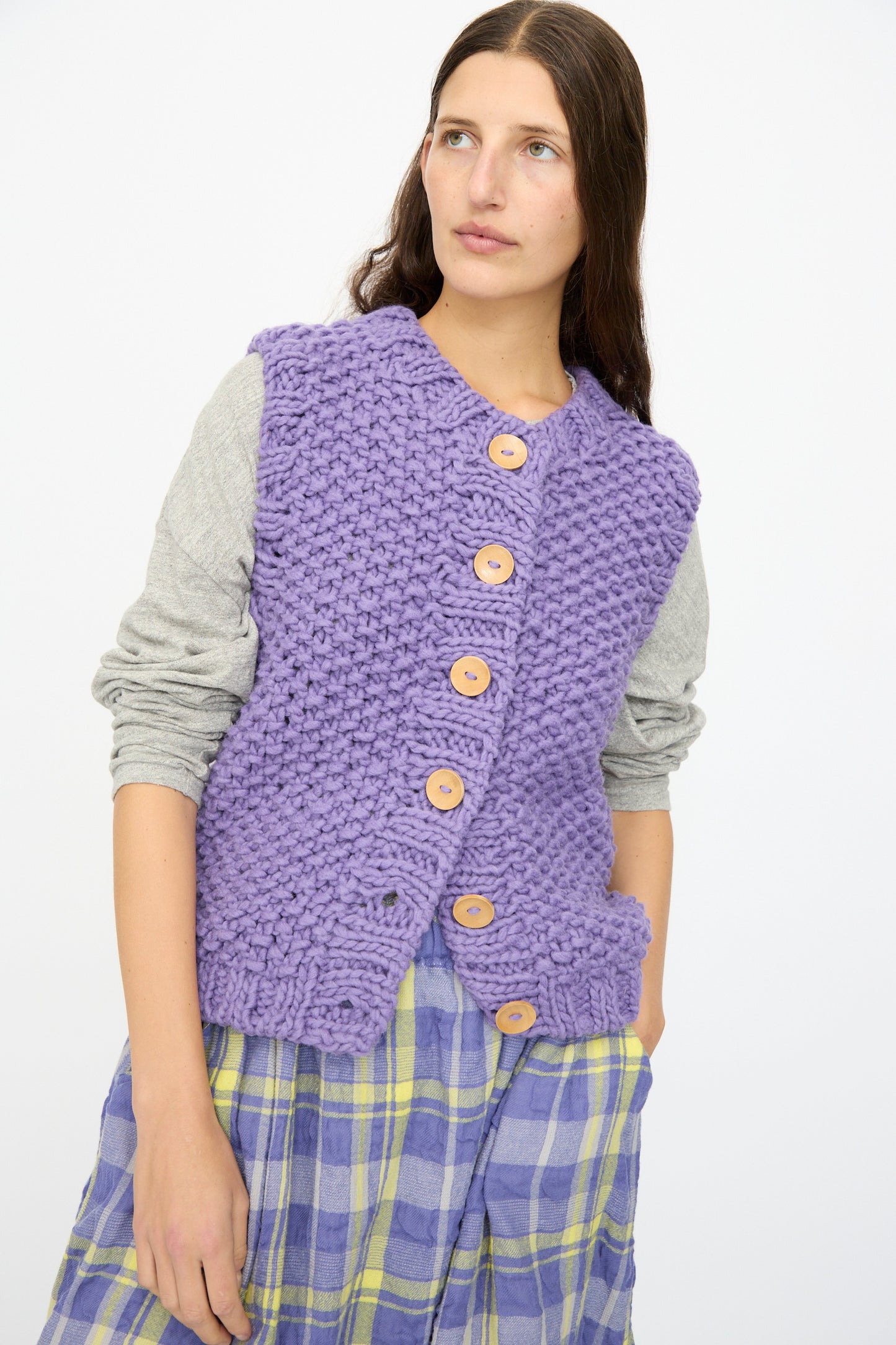 A woman wears the Ichi Antiquités Wool Peru Hand-knit Vest in Purple over a gray long-sleeve top and a yellow-and-blue plaid skirt, standing against a plain white background.