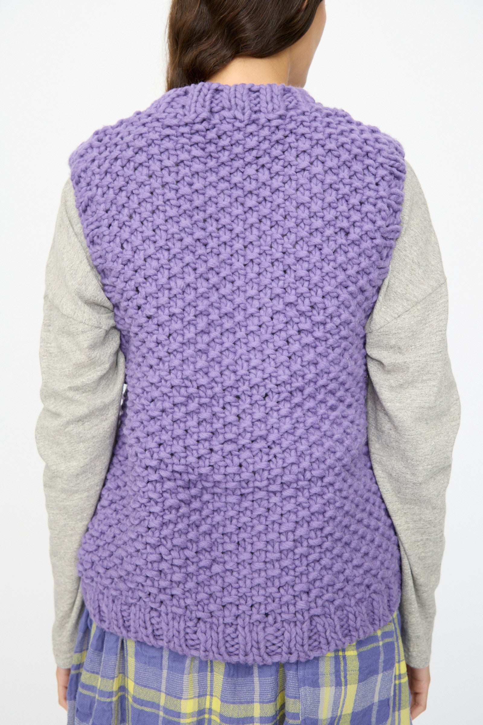 A person faces away, wearing the Ichi Antiquités Wool Peru Hand-knit Vest in Purple over a gray long-sleeve top and a blue, yellow, and gray plaid skirt.